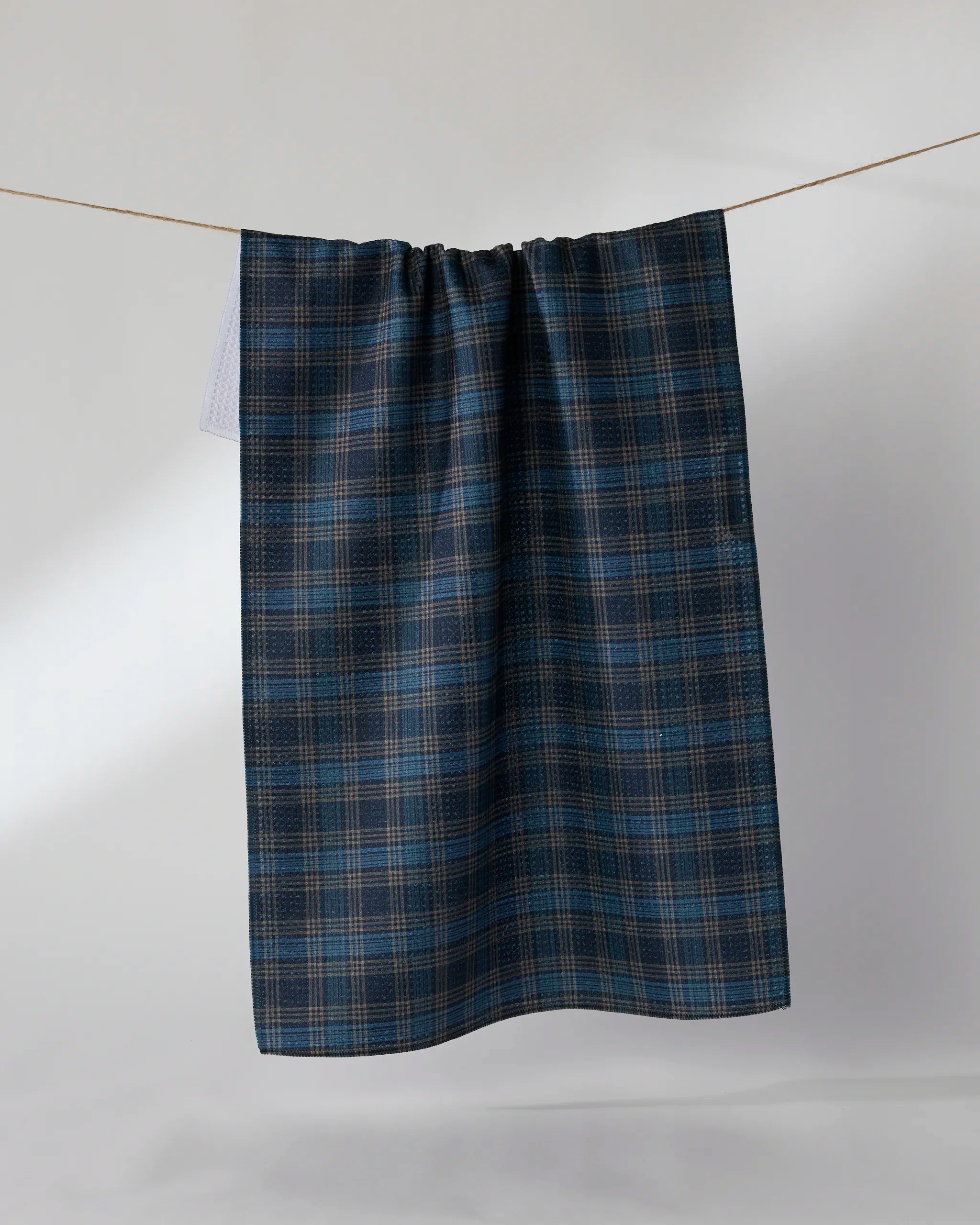 The Geometry Stonegate kitchen towel in blue and brown plaid hangs neatly over a clothesline against a light background, bringing an eco-friendly style to your daily routine.