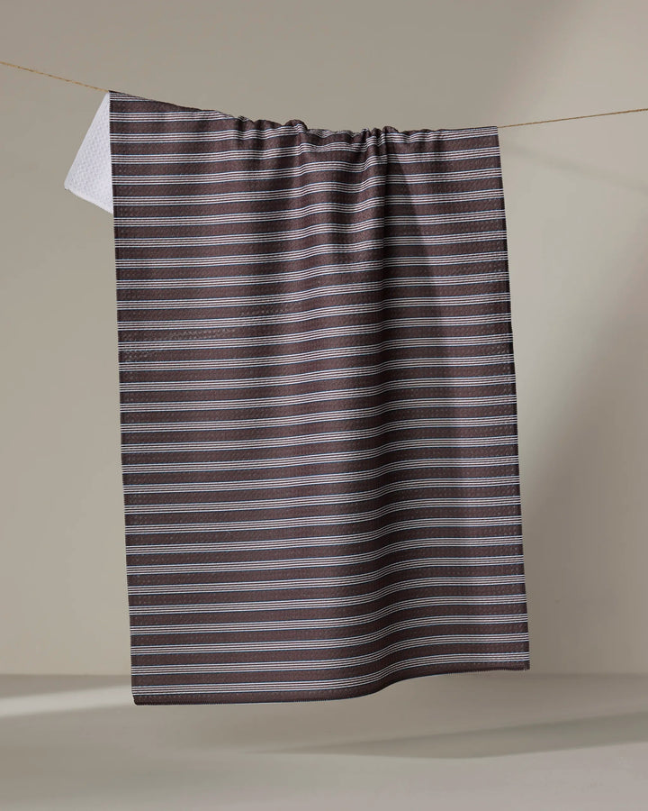 The Buy Guide’s Straight Across - Chocolate tea towel, featuring dark brown and gray stripes with a white inner side, hangs gathered on a clothesline against a plain beige background.