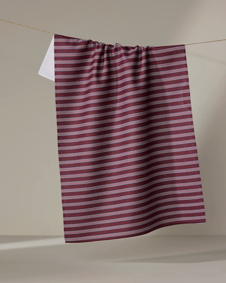 The Buy Guide’s Straight Across - Wine kitchen tea towel, crafted from recycled materials, in maroon and white stripes, hangs on a neutral background with one corner flipped to show the white underside.