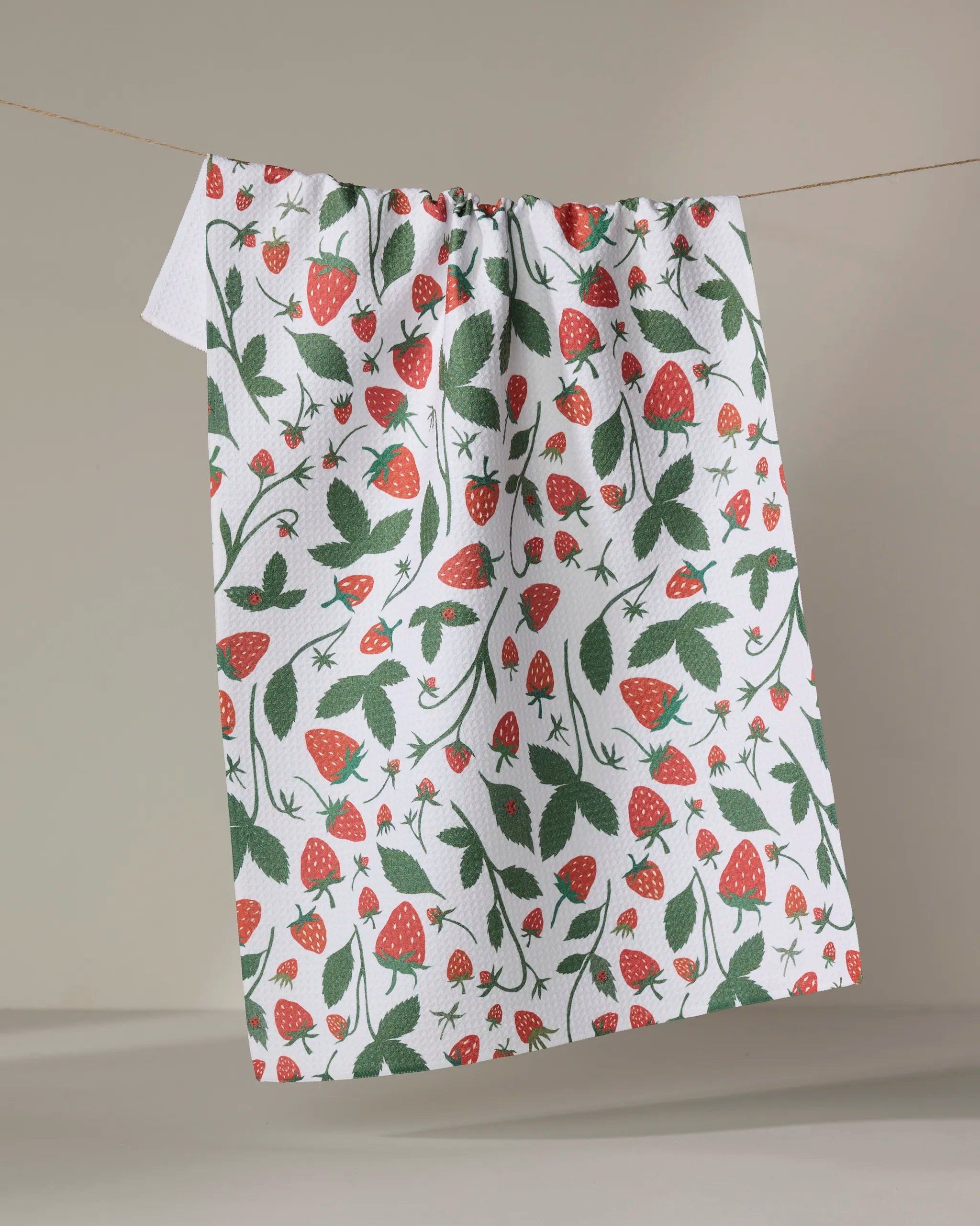 The "Strawberries" tea towel by Elana Gabrielle features a vibrant design of red strawberries, green leaves, and small flowers on white linen, displayed hanging against a plain beige background.