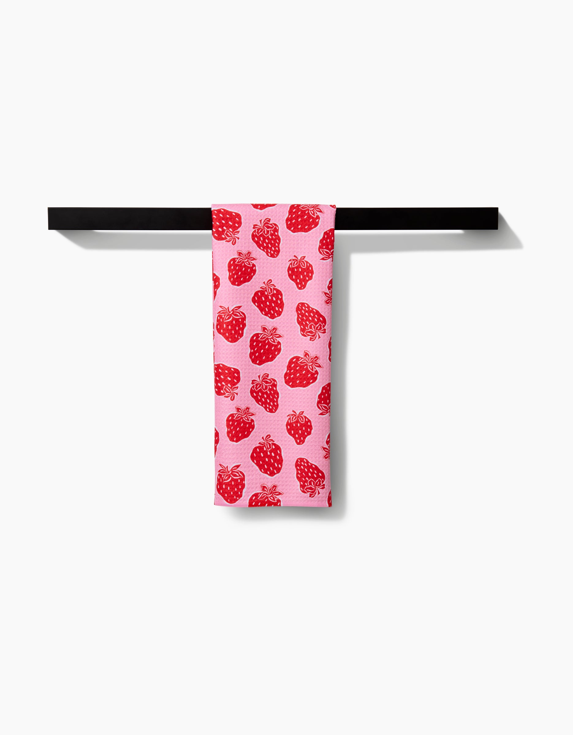The Strawberry Fields tea towel by Charlie Rowan Designs, in pink with a red strawberry pattern and soft Geoweave fabric, hangs neatly on a black bar against a white background.