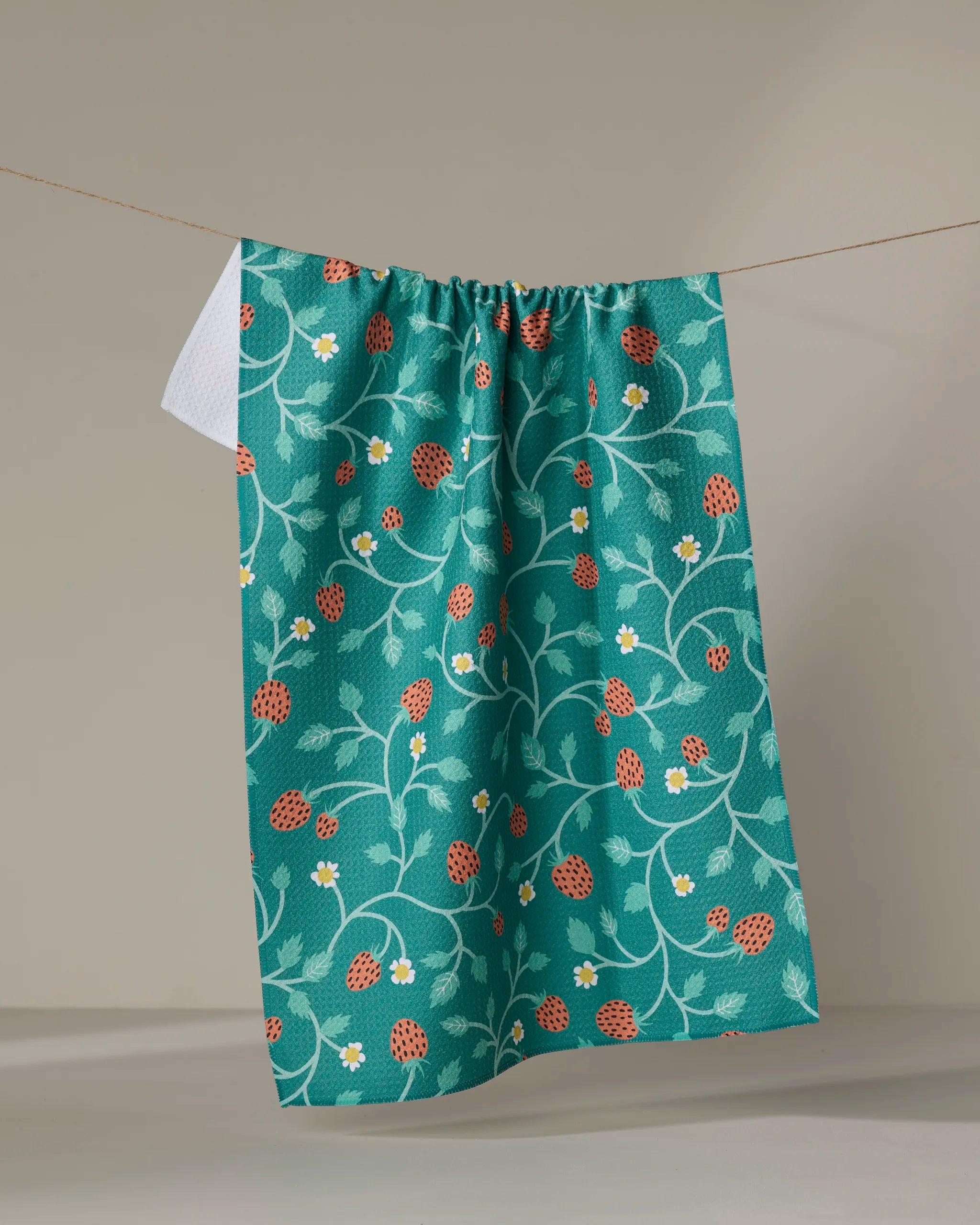 The "Strawberry Patch" kitchen tea towel by Megan Kampa, crafted from recycled materials and adorned with red strawberries, green vines, and white flowers, hangs against a neutral background.