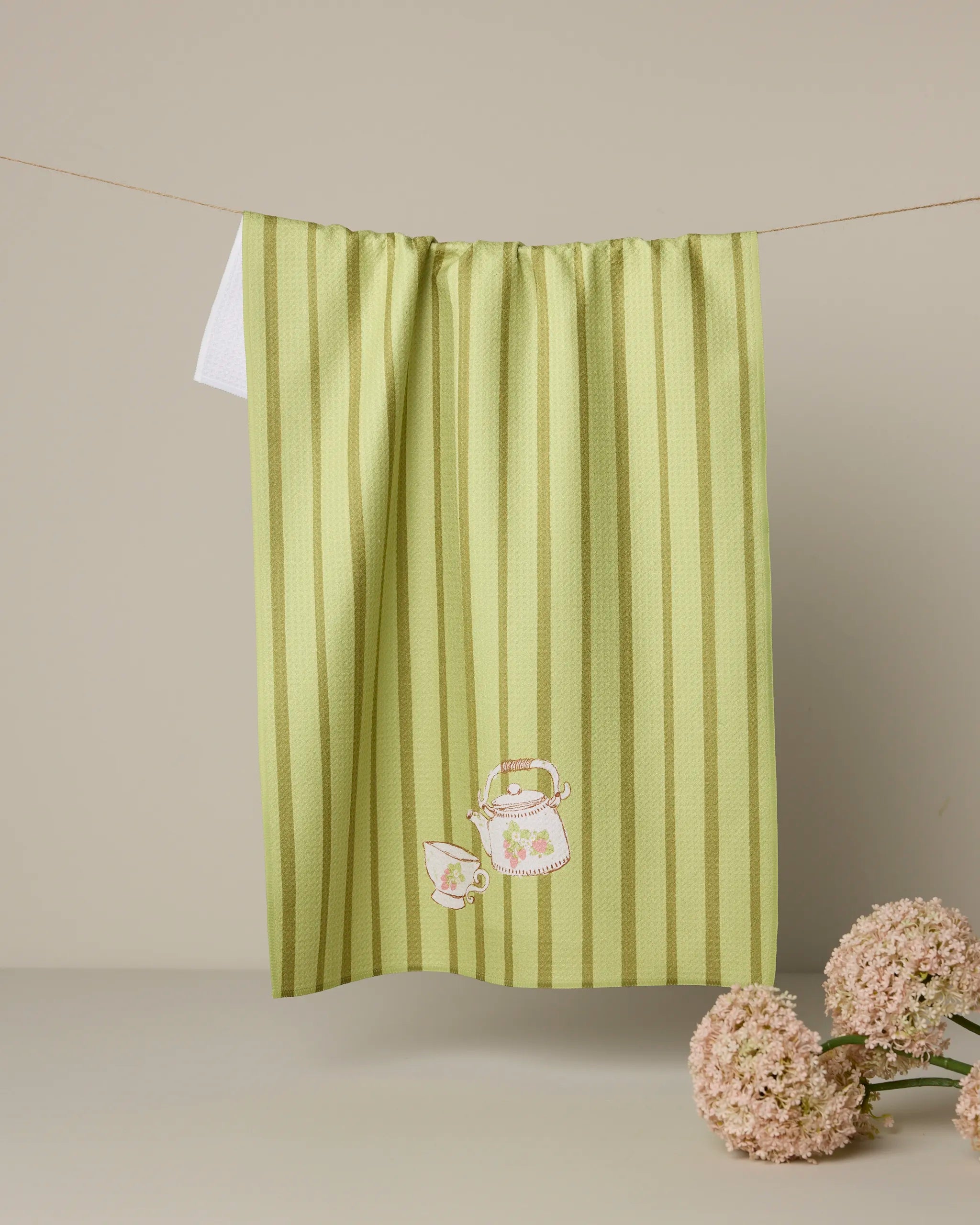 The Geometry "Strawberry Spring Green" tea towel, crafted from recycled materials and embroidered with a teapot and teacup, hangs on a line against a beige backdrop with pink hydrangeas at the bottom right.