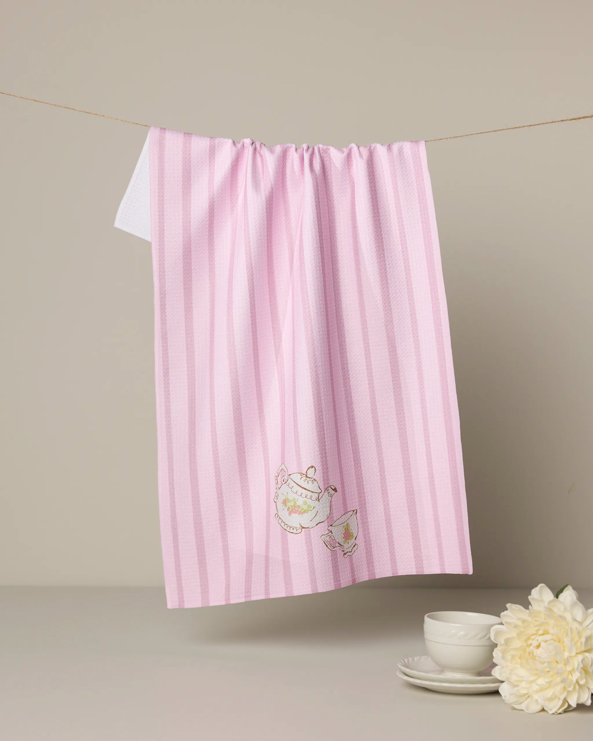The Geometry “Strawberry Spring” eco-friendly kitchen towel, featuring pink and white stripes with embroidered teapot and teacup designs, hangs on a line near white teacups, saucers, and a cream flower on a light surface.