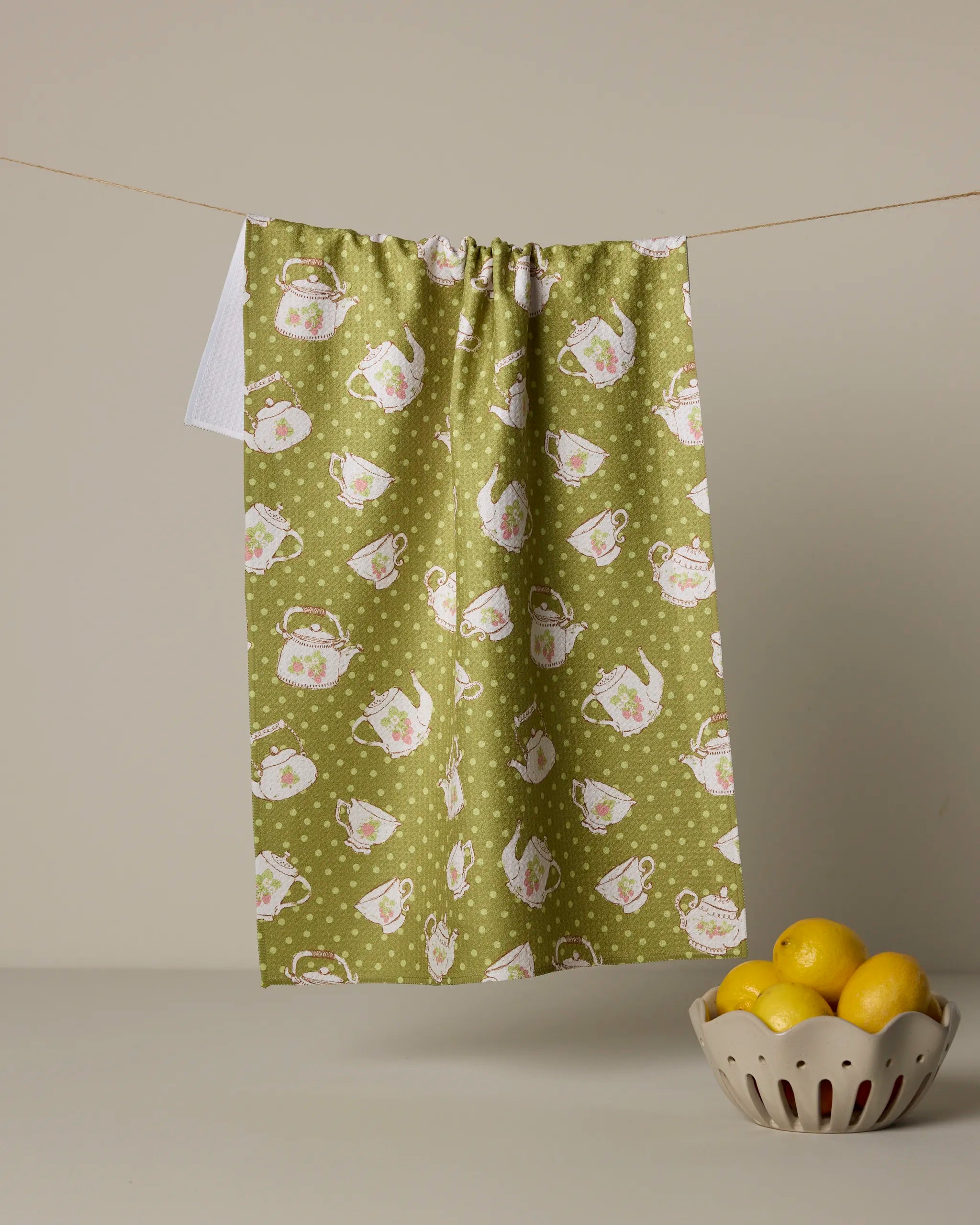 A Strawberry Tea Green kitchen towel by Geometry, featuring a teapot and polka dot design, hangs against a neutral wall. Below, a beige bowl of yellow lemons sits on a light surface.