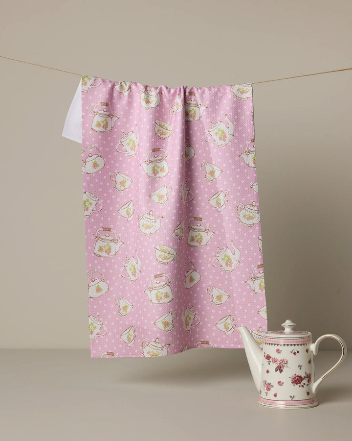 A Geometry "Strawberry Tea" pink kitchen towel with teapots and cups hangs on a clothesline above a white ceramic teapot with pink floral patterns, set on a light surface against a beige background.