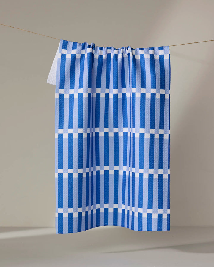 The Buy Guide’s Stripe Block - Blues is an absorbent kitchen towel made from recycled materials, featuring blue and white grid stripes. It hangs on a clothesline against a light background.