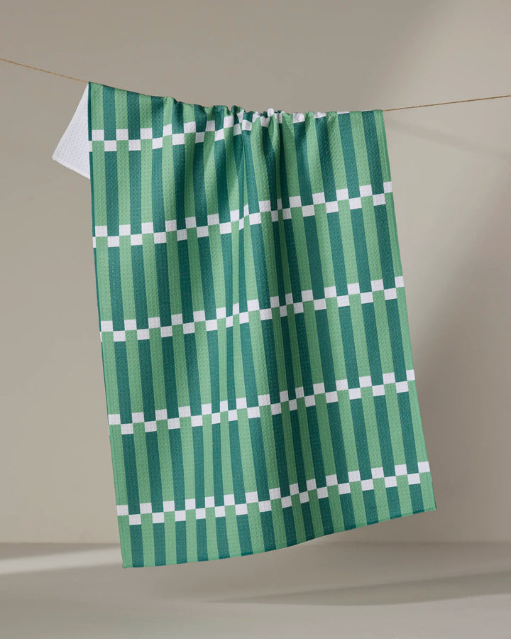 The Stripe Block - Greens tea towel by The Buy Guide hangs on a line against a neutral background, showcasing its green and white geometric pattern with vertical stripes and evenly spaced horizontal white lines.