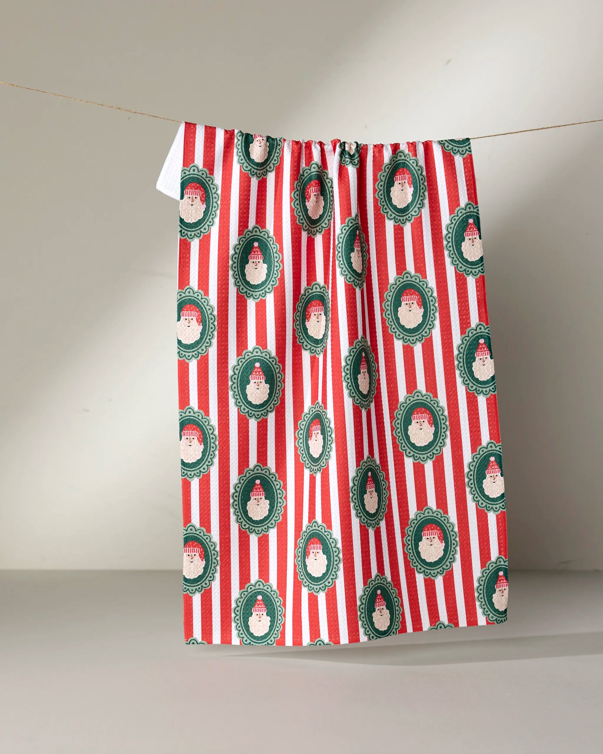 The Striped Santa tea towel by Charlie Rowan Designs features red and white stripes with repeating green oval Santa faces, made from recycled materials, and is shown hanging on a thin string against a neutral background.