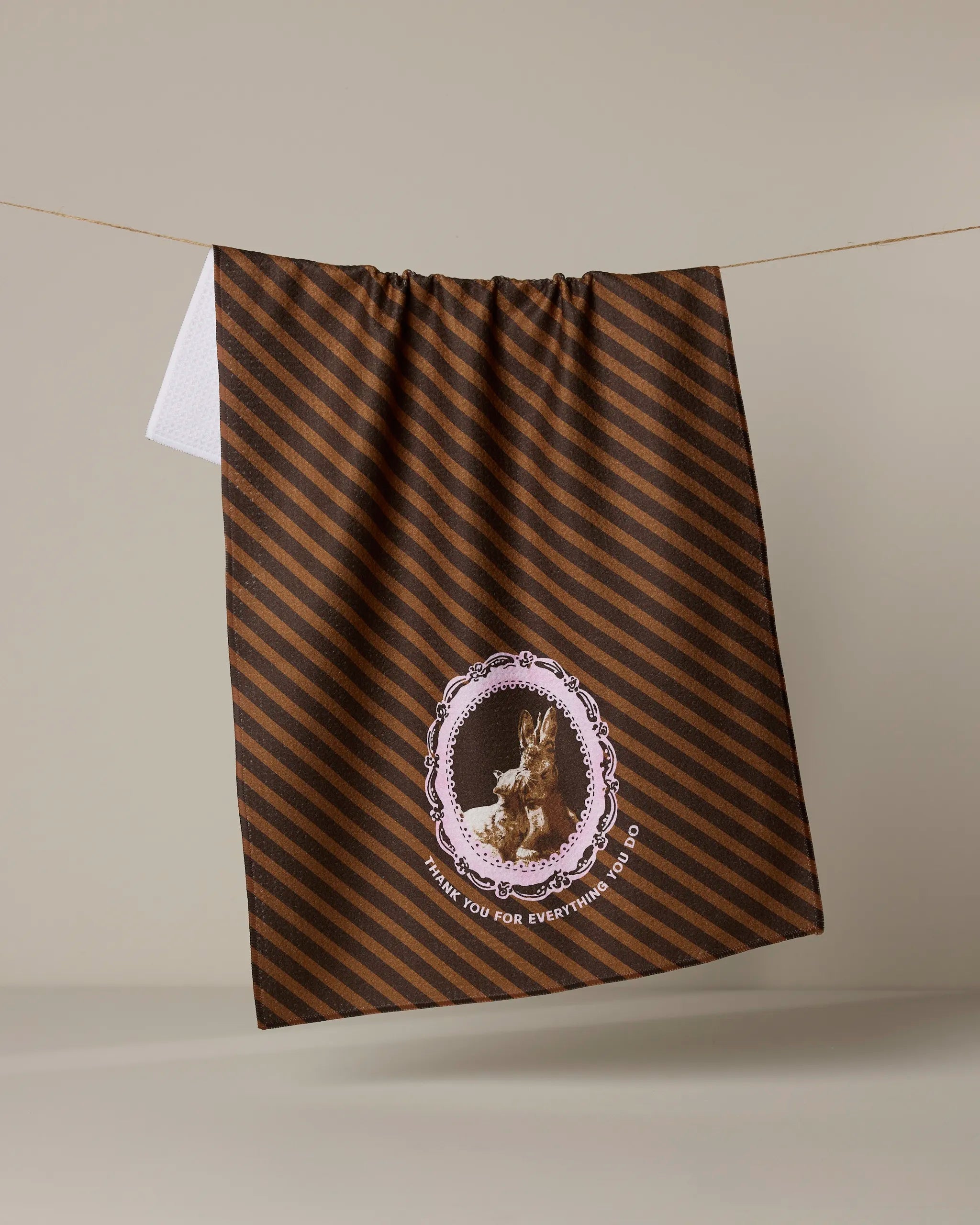 A brown Geometry “Strong and Caring” kitchen tea towel with diagonal dark stripes hangs on a line. Made from recycled materials, it features a framed rabbit illustration and “THANK YOU FOR EVERYTHING YOU DO” underneath.