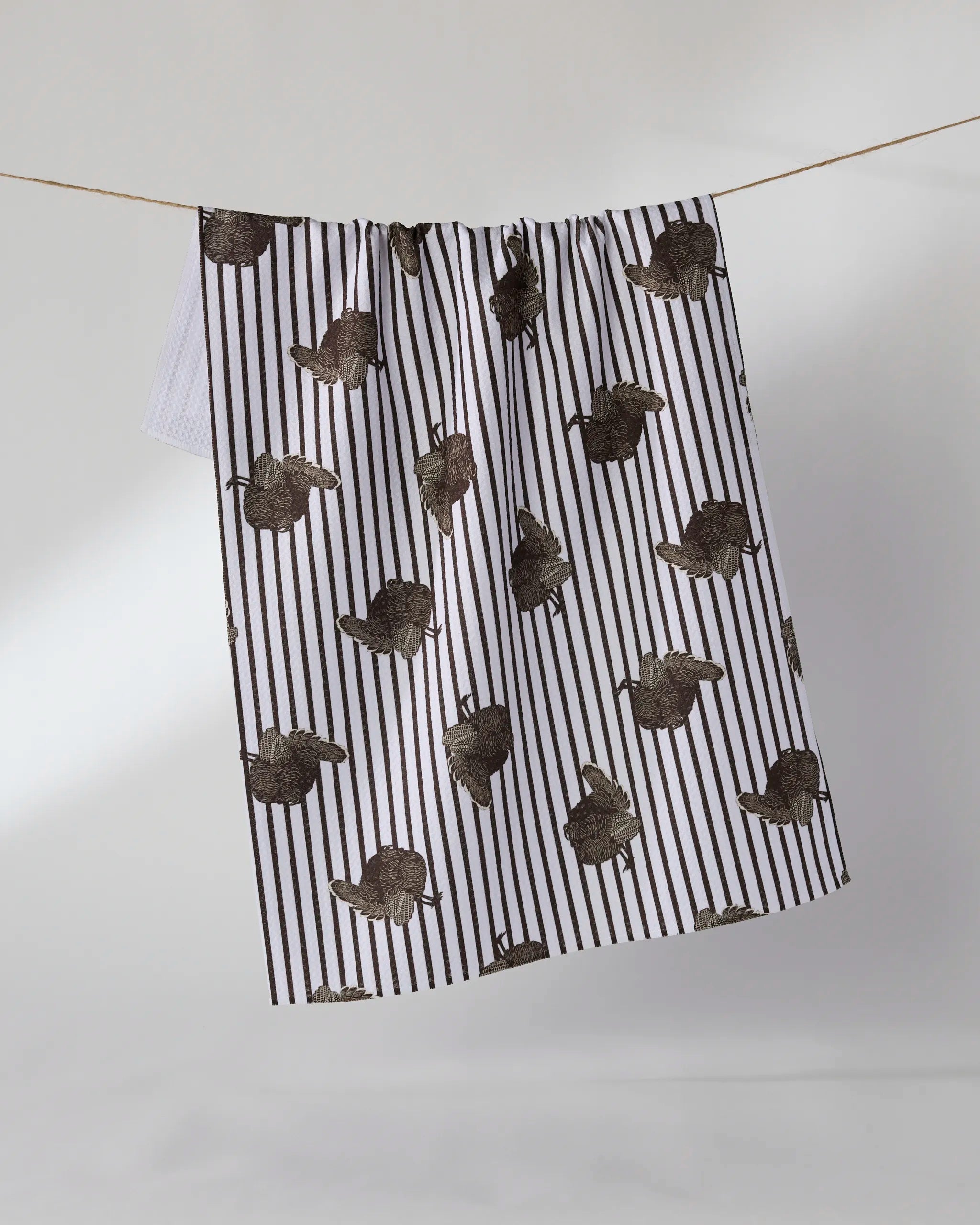 The Strut tea towel by Geometry, featuring black vertical stripes and brown leaf patterns on white, is made from quick-drying recycled materials and hangs on a clothesline against a light background.