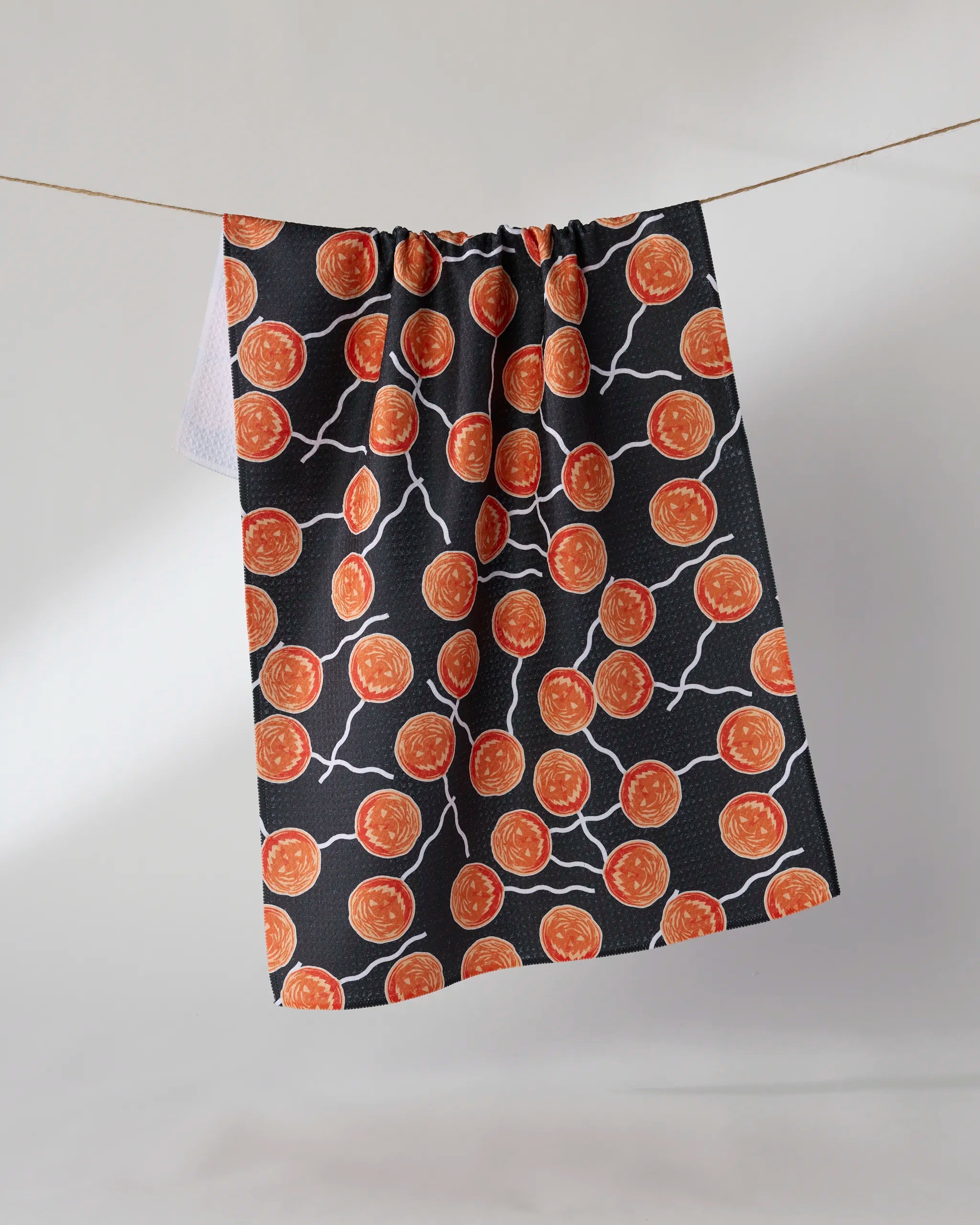 The Geometry "Sugar Shock" black tea towel, crafted from recycled materials and featuring orange circles with white lines, hangs on a string against a plain light background.