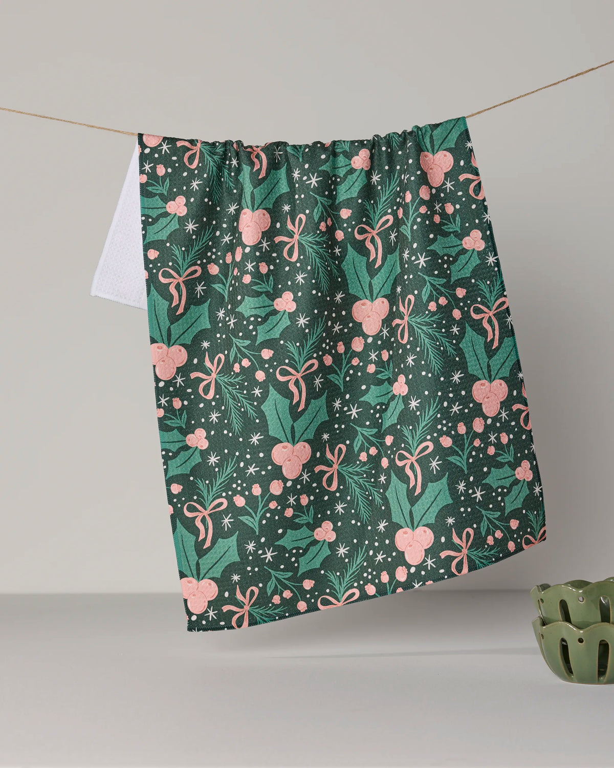 The Sugarplum Florals tea towel by Geometry is crafted from recycled materials and displays a cheerful green background adorned with pink bows, holly leaves, berries, and white snowflakes as it hangs on a line above a gray surface.