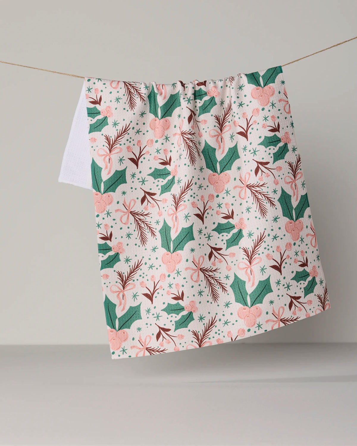 A Geometry kitchen towel in the “Sugarplum Florals” design, featuring a festive pink, green, and white holiday pattern with birds, hearts, stars, and leaves, hangs on a clothesline against a plain light gray background.