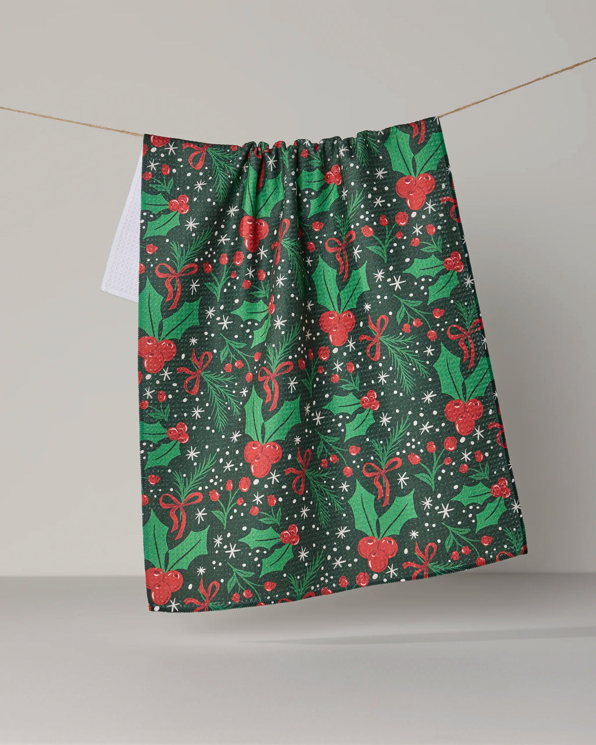 A festive tea towel from Geometry, the Sugarplum Florals, featuring a vibrant holiday design, hangs on a string against a plain gray background. Crafted from durable Geoweave fabric and made with recycled materials, this eco-friendly cloth brings seasonal cheer to any kitchen.