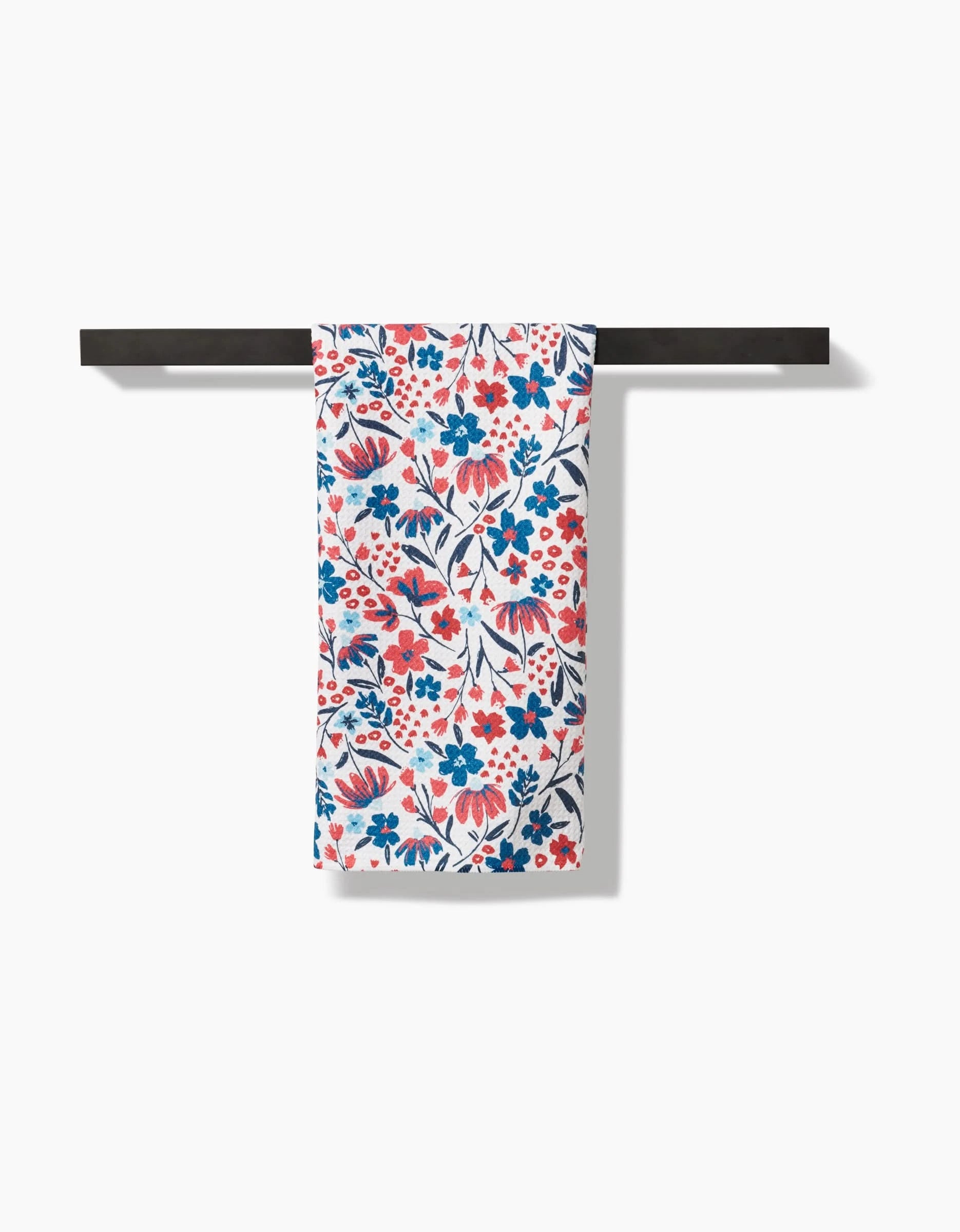The Summer Bloom Tea Towel by Hufton Studio features a vibrant red, blue, and orange floral pattern. Made from recycled materials, it brings sustainable charm to your kitchen, shown here against a crisp white background on a black rod.