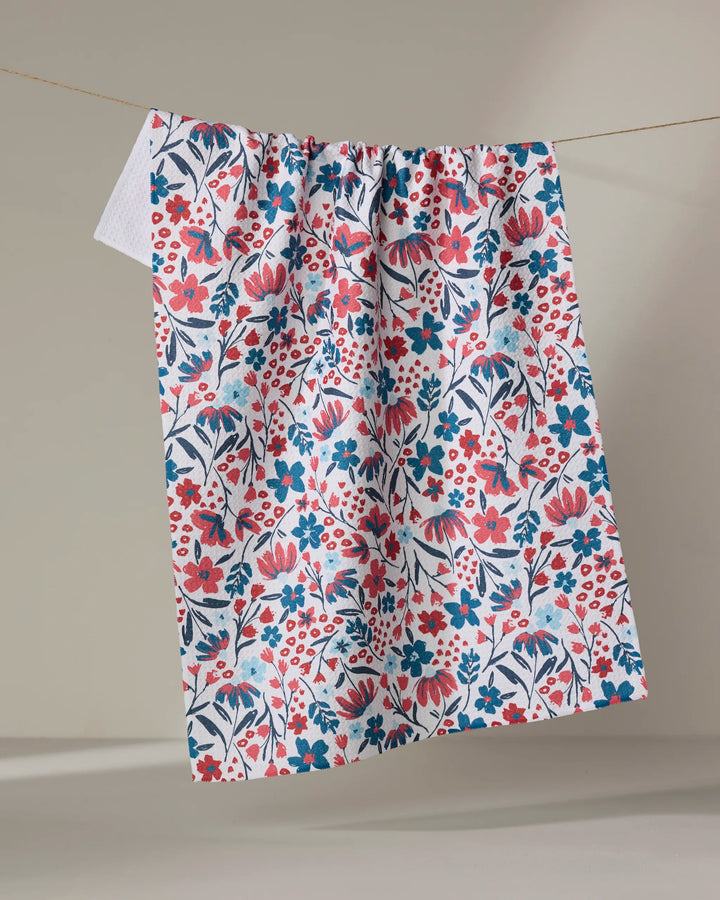 The Summer Bloom kitchen towel by Hufton Studio, crafted from recycled materials, features a colorful red, pink, and blue floral pattern on white and hangs on a string against a neutral background.