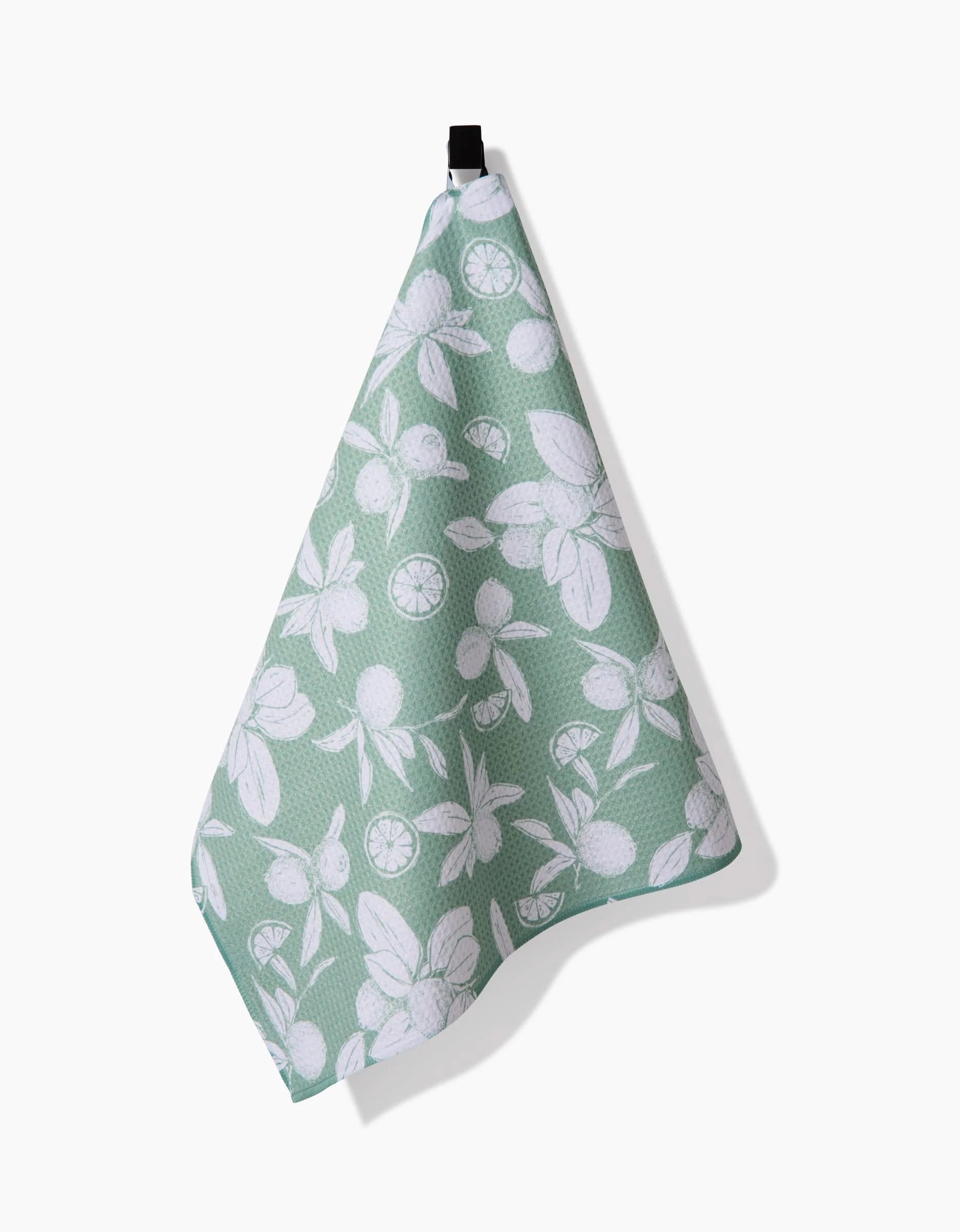 The Hufton Studio Summer Limes tea towel, in light green with a white citrus fruit and leaf pattern, crafted from recycled materials, hangs by a black loop against a white background.