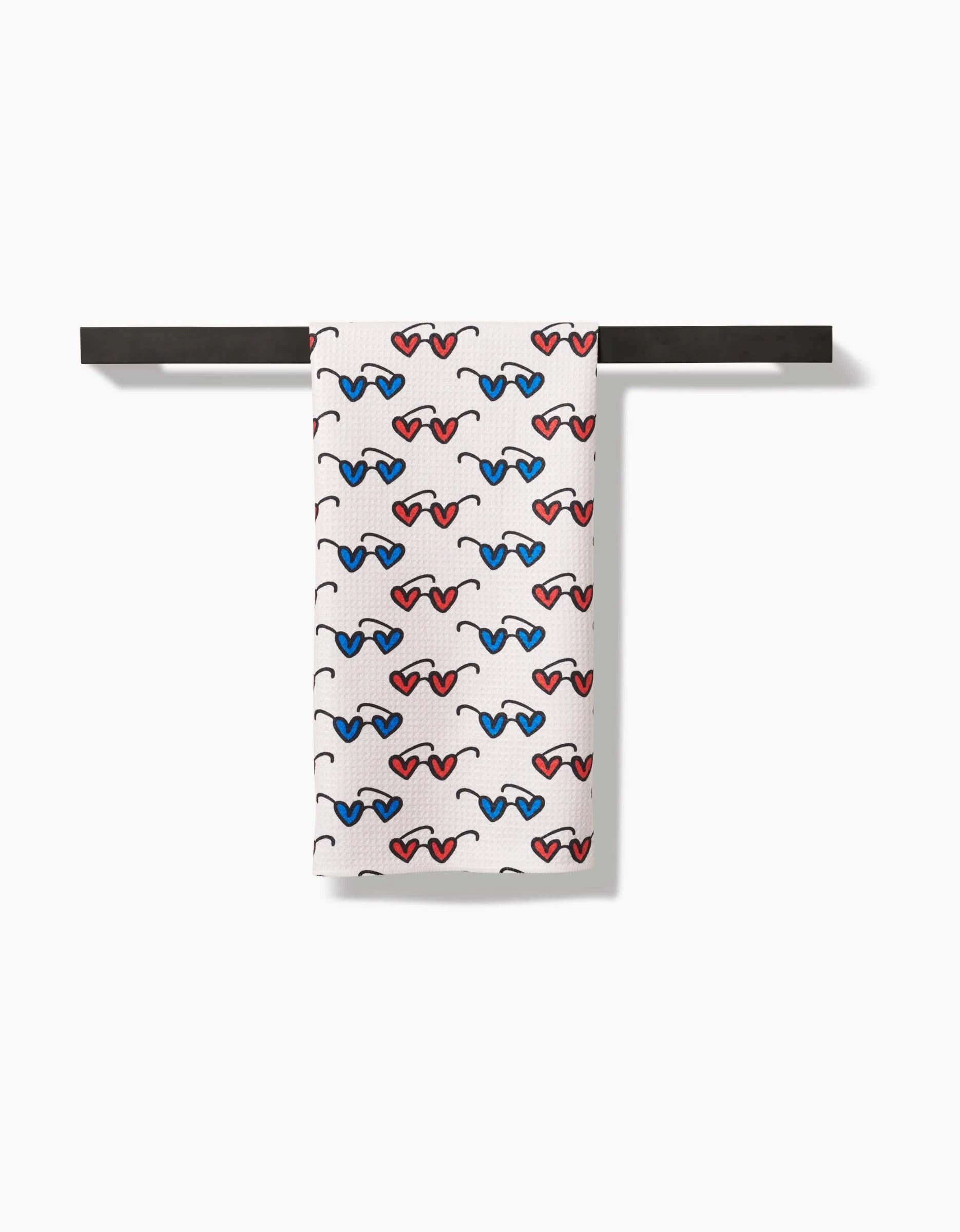 The Kayla Ann Summer Sunglasses tea towel, made from recycled materials, features a repeating red, blue, and black sunglasses pattern. Shown neatly draped over a sleek black bar against a plain white background.