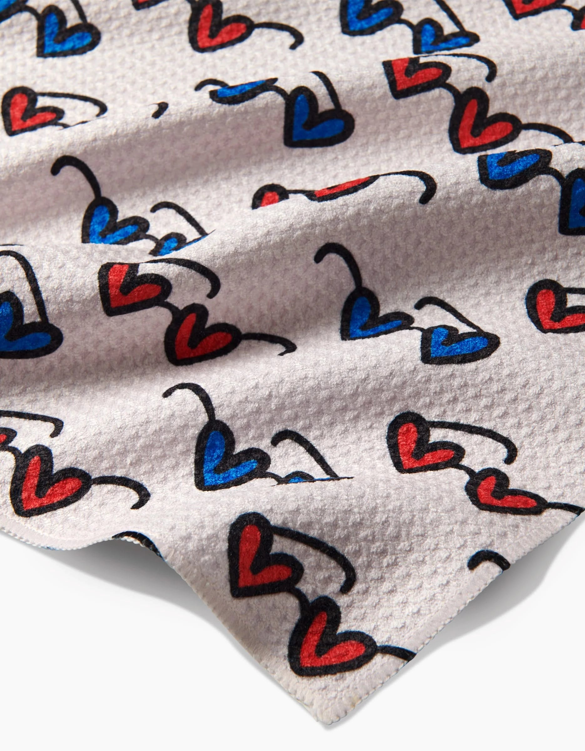 Summer Sunglasses by Kayla Ann feature a playful design with red and blue heart shapes outlined in black, set against beige recycled material with textured fabric folds and shadows.