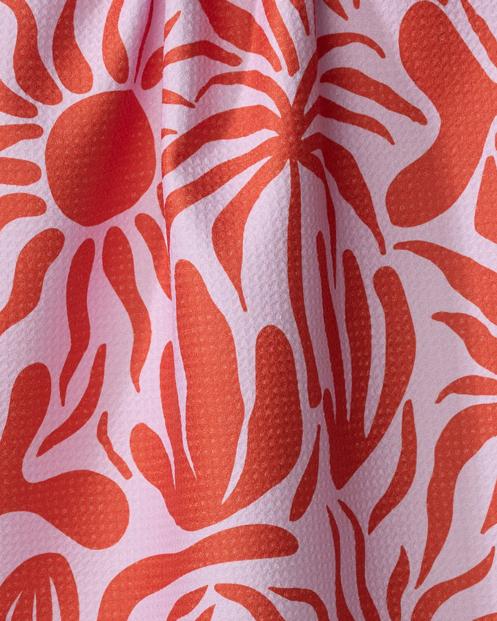 A close-up of the “Summer Sunset” dish towel by Marina Solodka reveals pink textured fabric with abstract red sunburst and flowing patterns. The absorbent material is softly gathered, creating gentle folds and shadows.