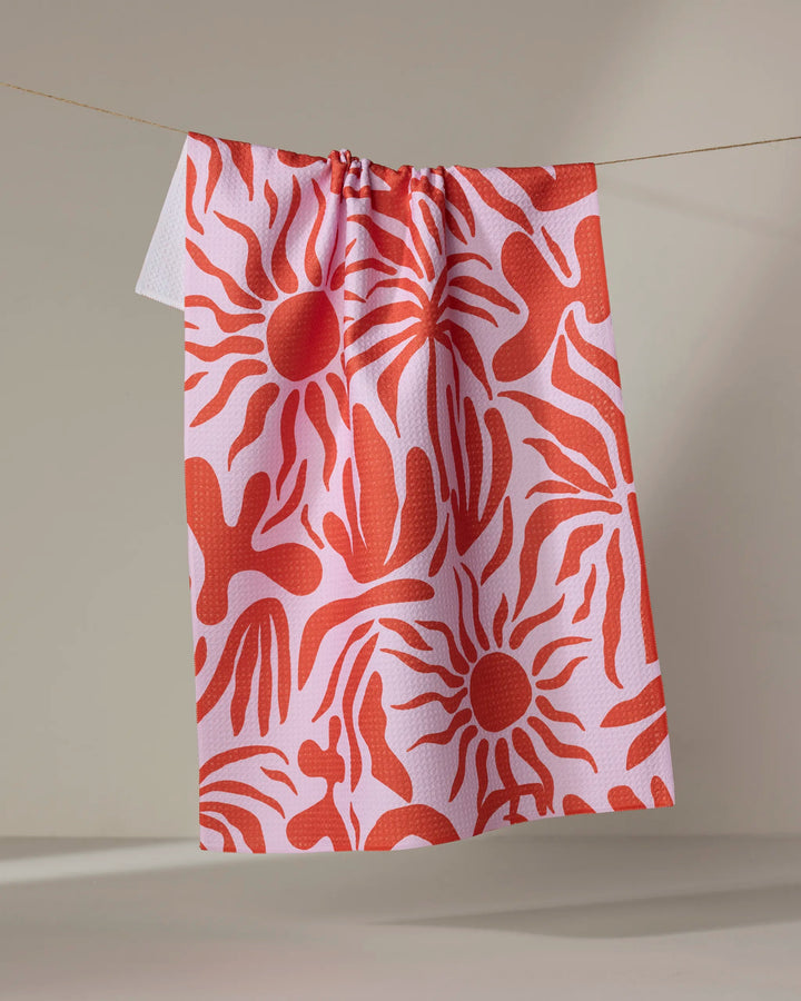 The "Summer Sunset" recycled kitchen towel by Marina Solodka, featuring bold red sunburst and hand-like patterns on pink, hangs on a string against a beige background.