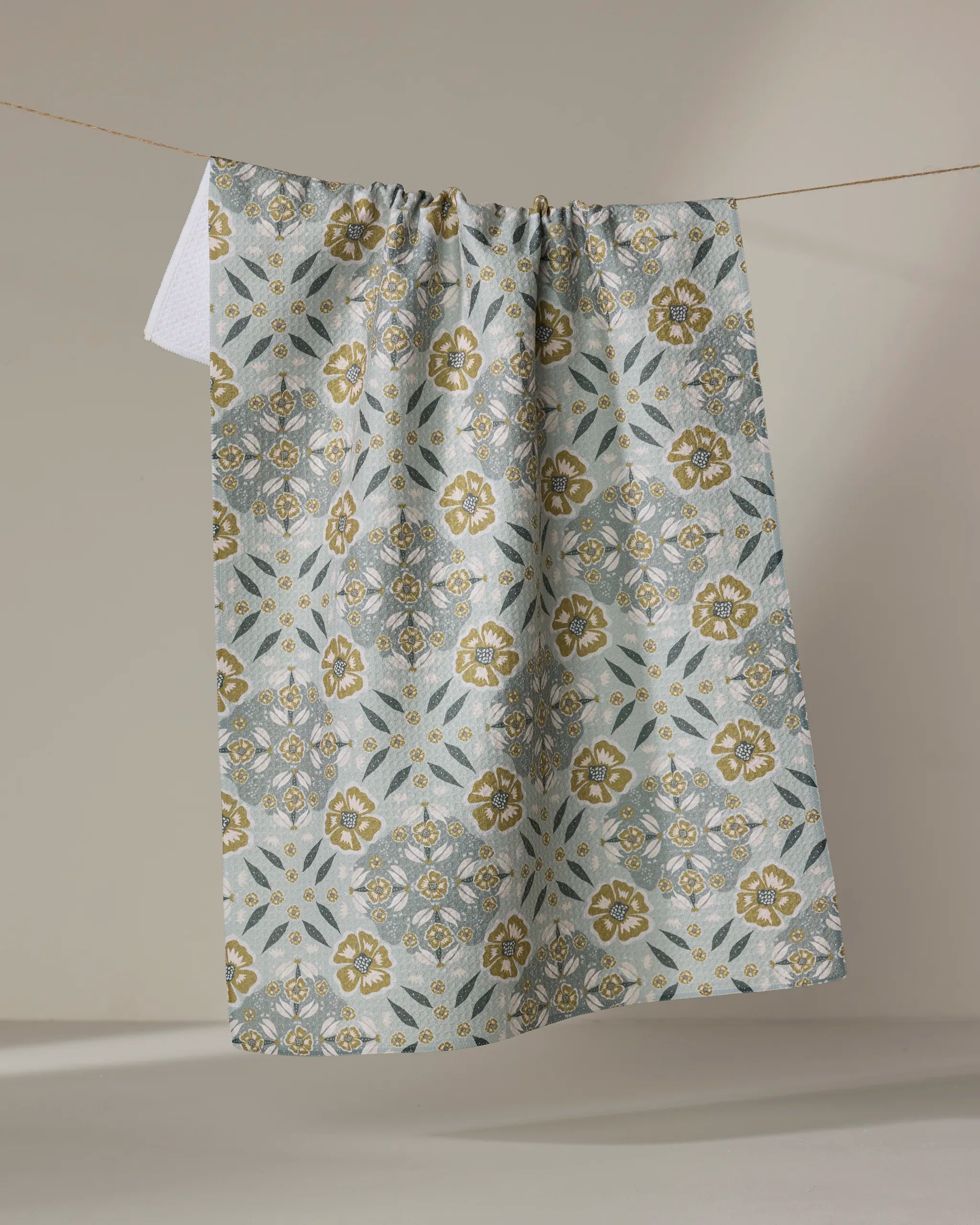 The Ash Cascade Summit Check recycled kitchen towel with gold and light blue floral patterns hangs on a clothesline against a neutral background.