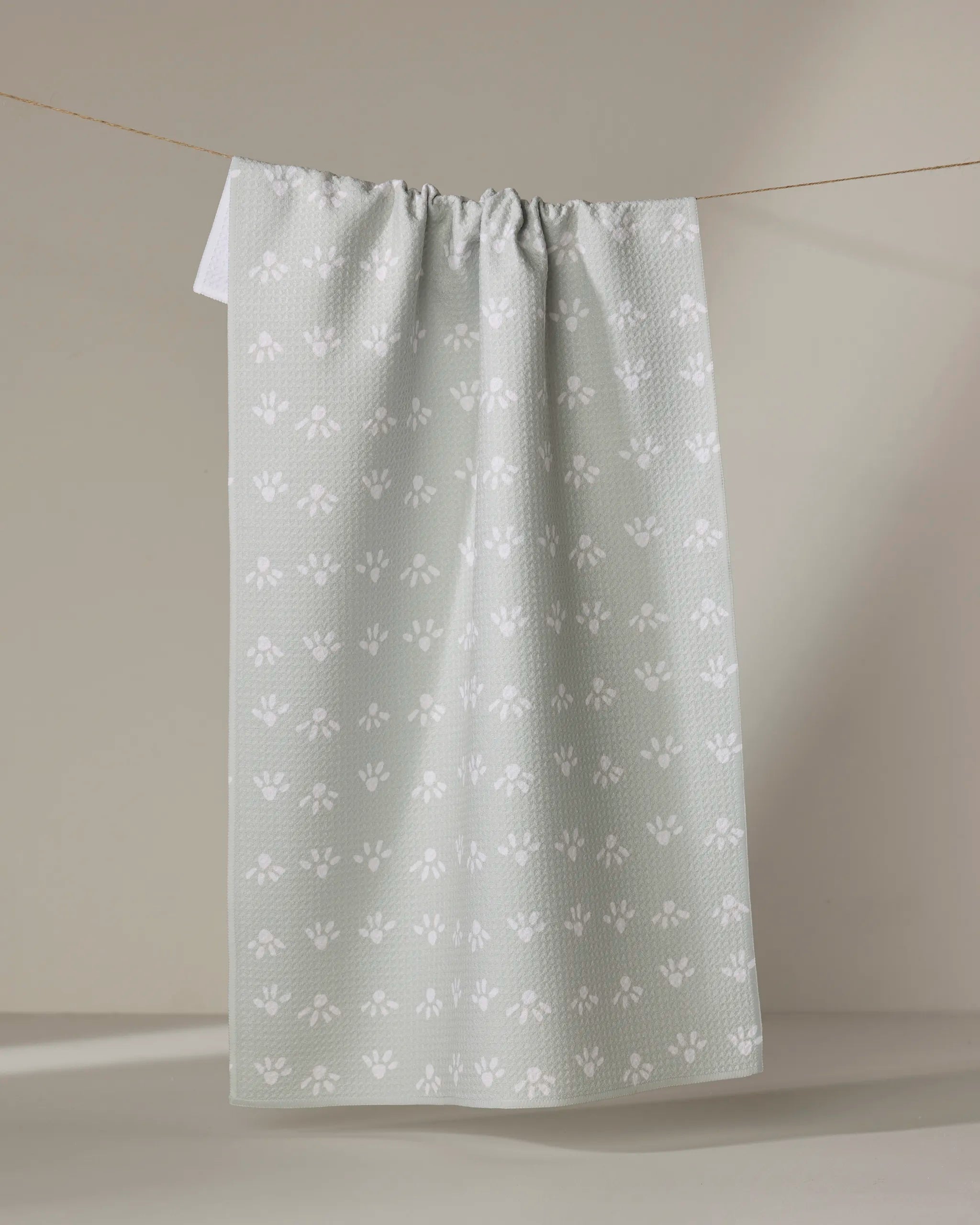 The "Sun Bursts" kitchen tea towel by Anee Shah, crafted from recycled materials in soft green with a white floral pattern, hangs from a string against a neutral beige background.