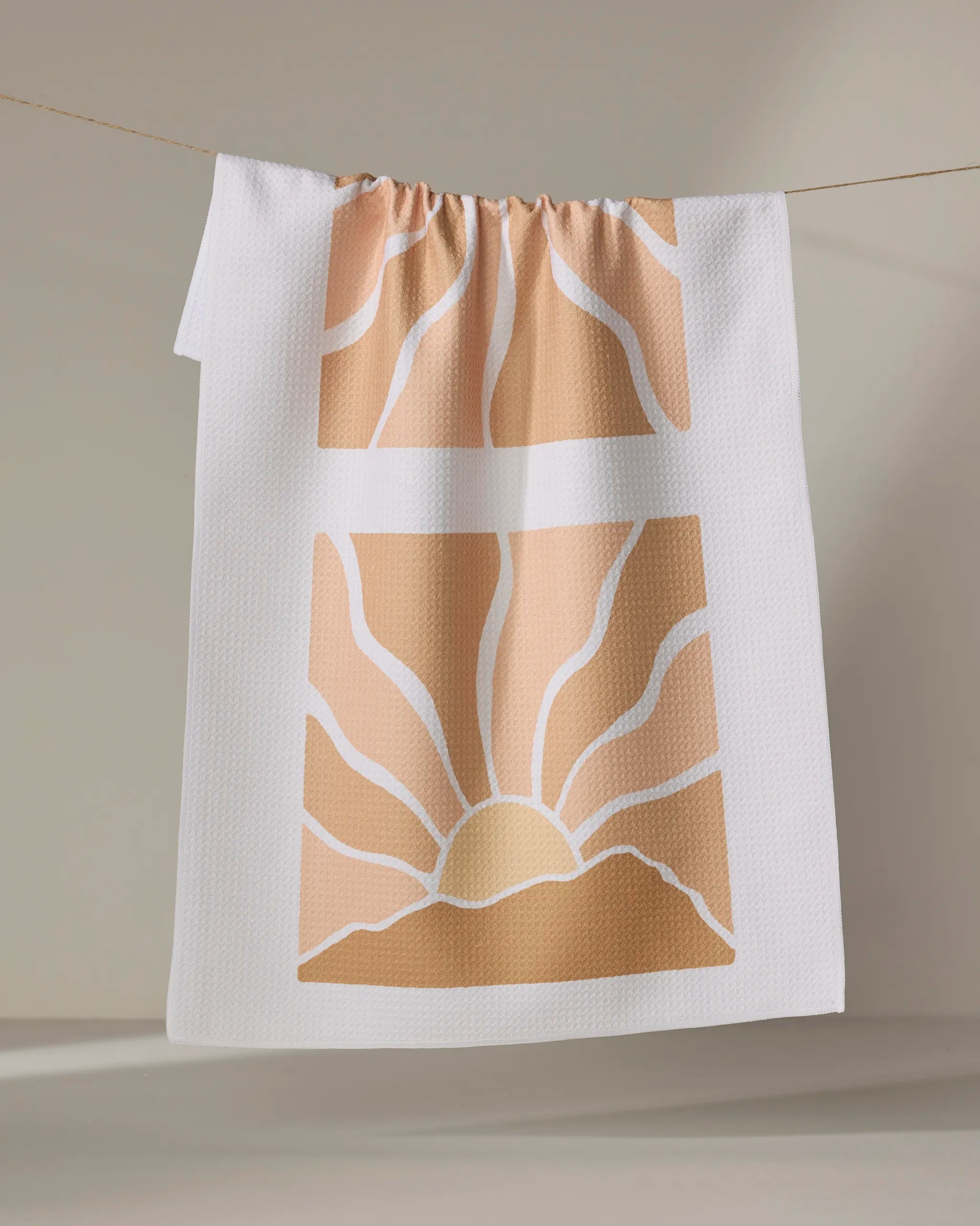 The Sun Rays kitchen towel by Graphics and Grain features a minimalist rising sun with rays and hills in warm tan and peach on white textured fabric. Crafted from recycled materials, it brings eco-friendly style to your kitchen.