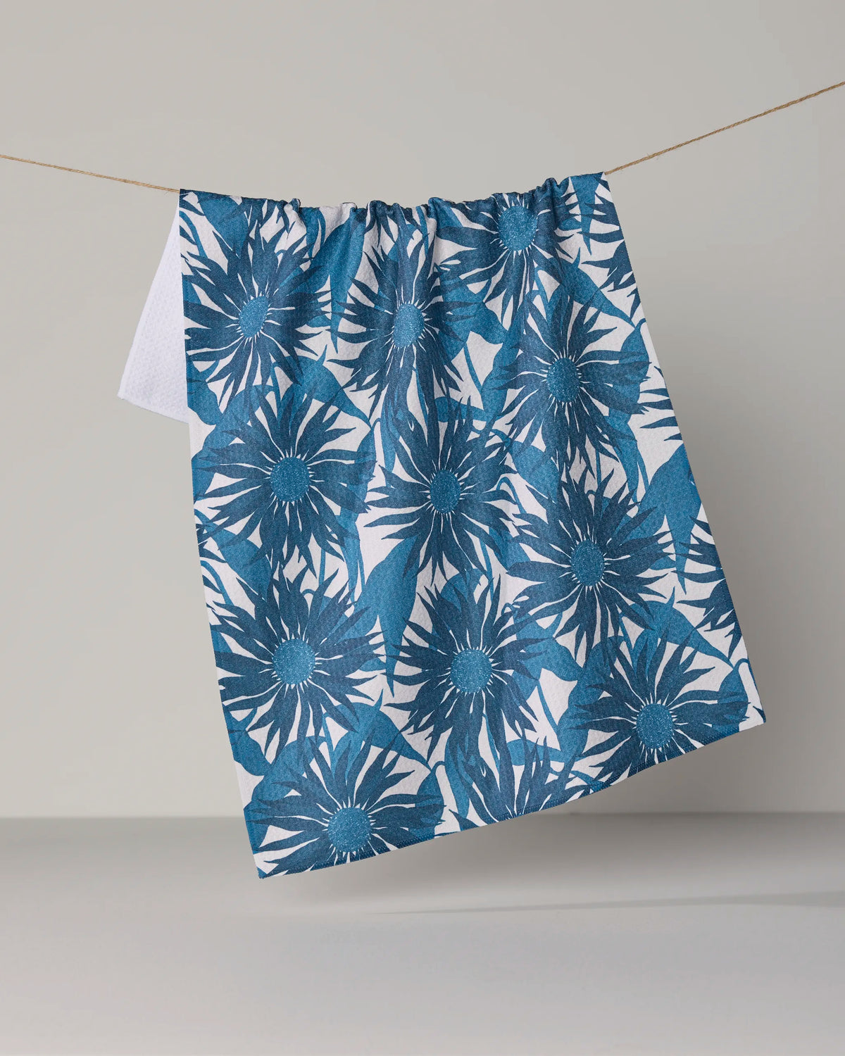 A Geometry Sundara Frost towel, featuring a blue floral pattern on ultra-absorbent white fabric, hangs on a string against a plain, light-colored background.