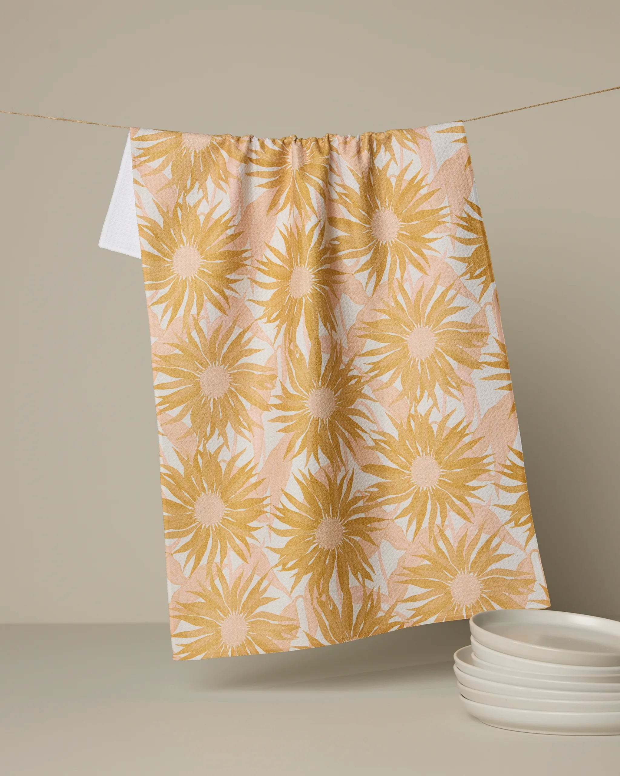 The Geometry Sundara kitchen tea towel, featuring a yellow sunflower print and crafted from recycled materials, hangs above a stack of white plates, all set against a neutral background.