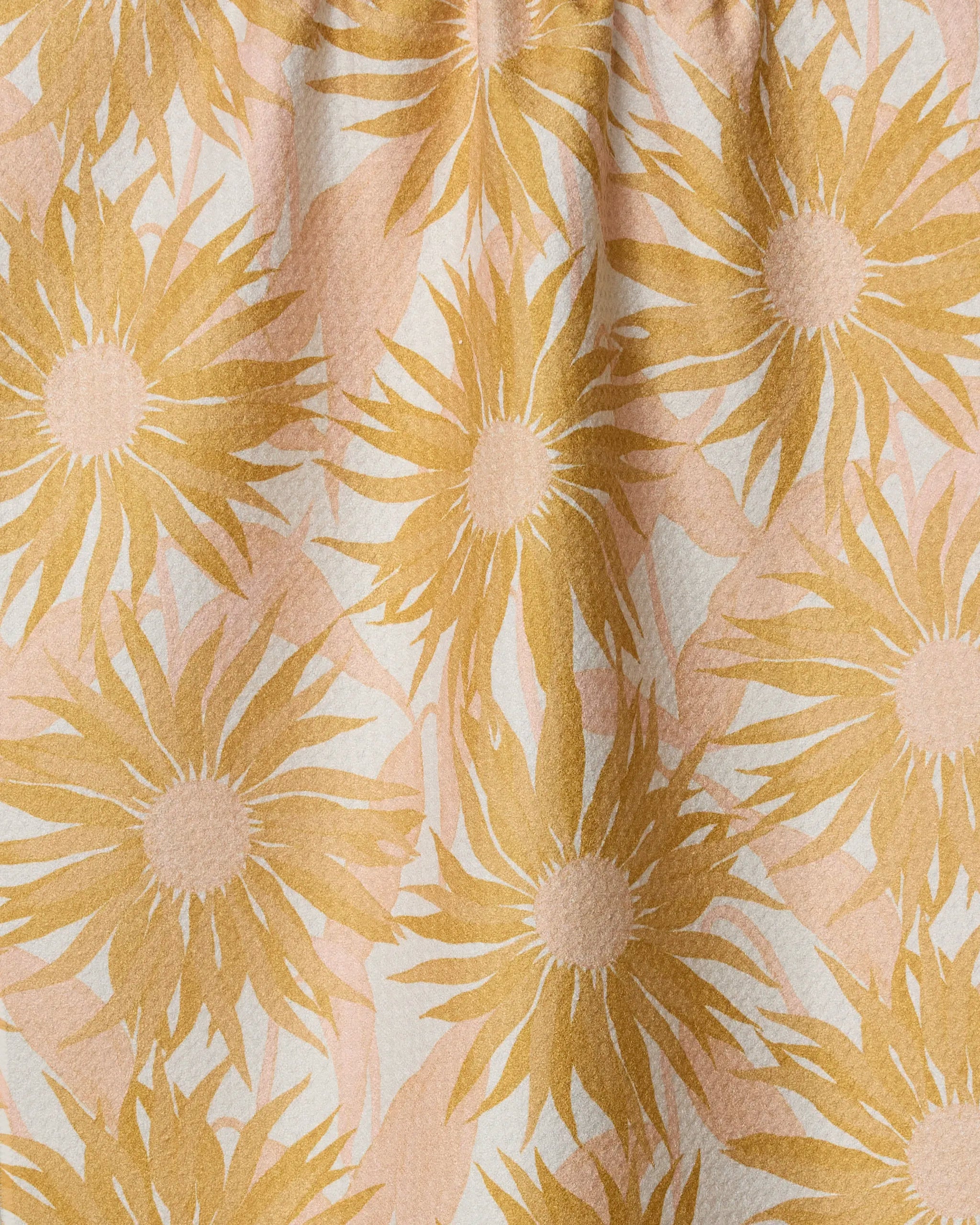 The Sundara kitchen towel by Geometry features a repeating pattern of large, golden-yellow sunflowers and light beige leaves on a white background. Made from recycled materials, its subtle texture and soft folds add visual interest.