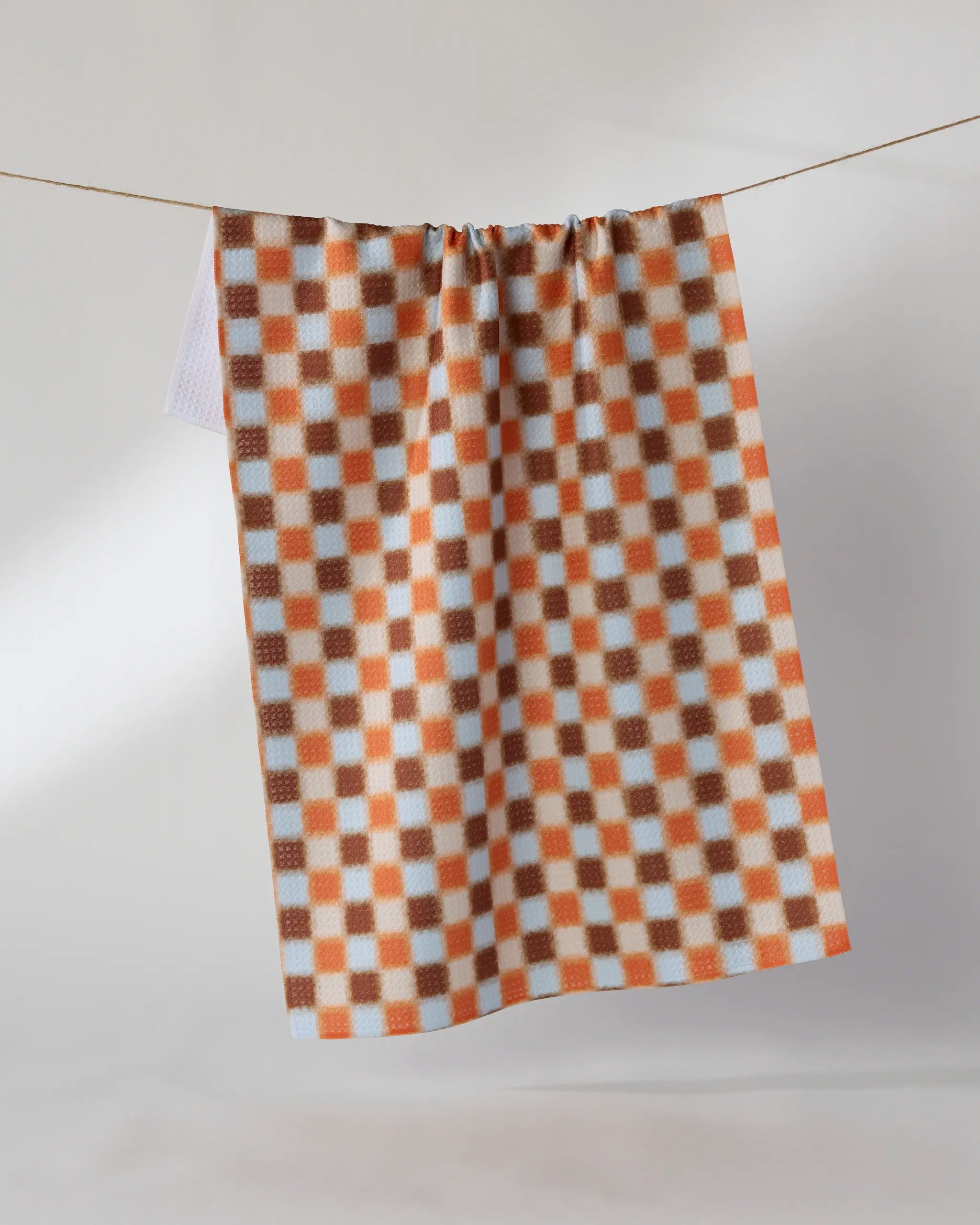 The Geometry Sunday Supper tea towel, featuring an orange and white checkered pattern, hangs on a clothesline against a plain light background. This ultra-absorbent kitchen towel combines style and function.