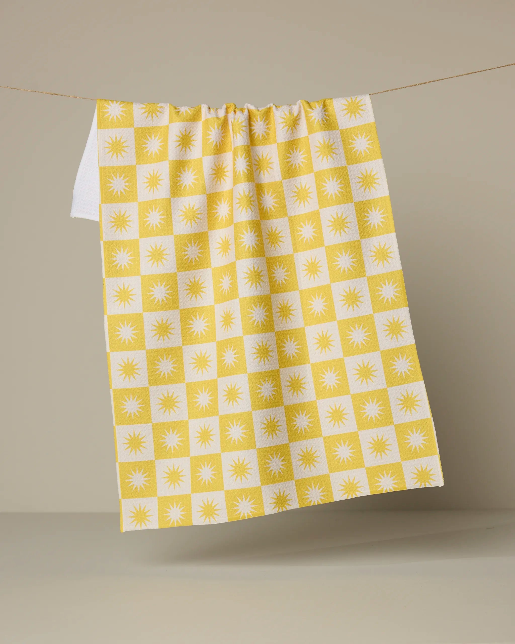 The Sundial tea towel by Geometry, featuring a yellow and white checkered sunburst pattern and made from recycled materials, hangs on a clothesline against a neutral beige background.