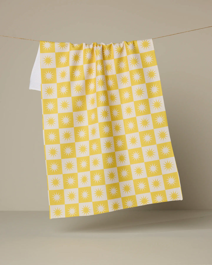 The Sundial tea towel by Geometry, featuring a yellow and white checkered sunburst pattern and made from recycled materials, hangs on a clothesline against a neutral beige background.