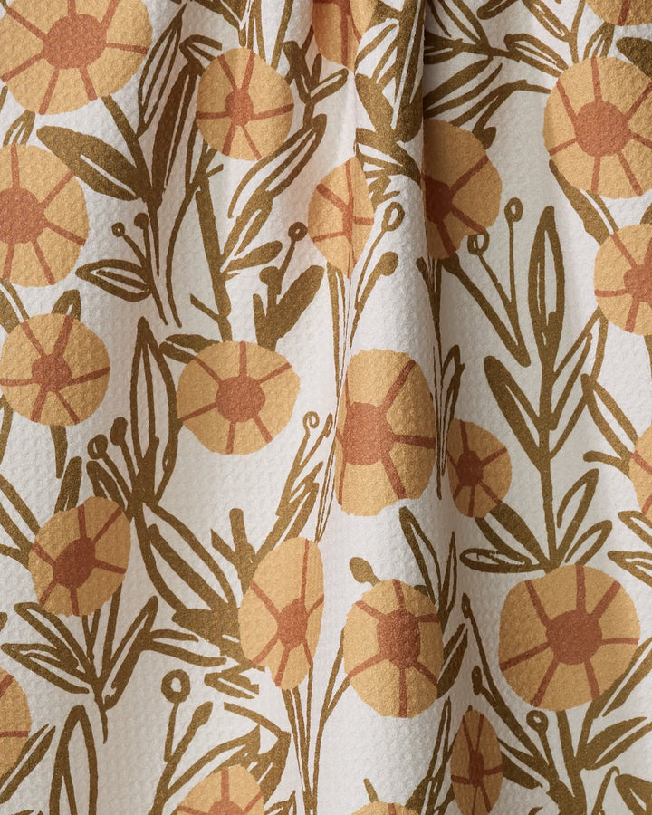 A close-up of the "Sunflower Fields" tea towel by Janna Sue Design shows yellow and orange floral patterns with greenish-brown leaves on a textured white background. This ultra-absorbent towel is softly folded.