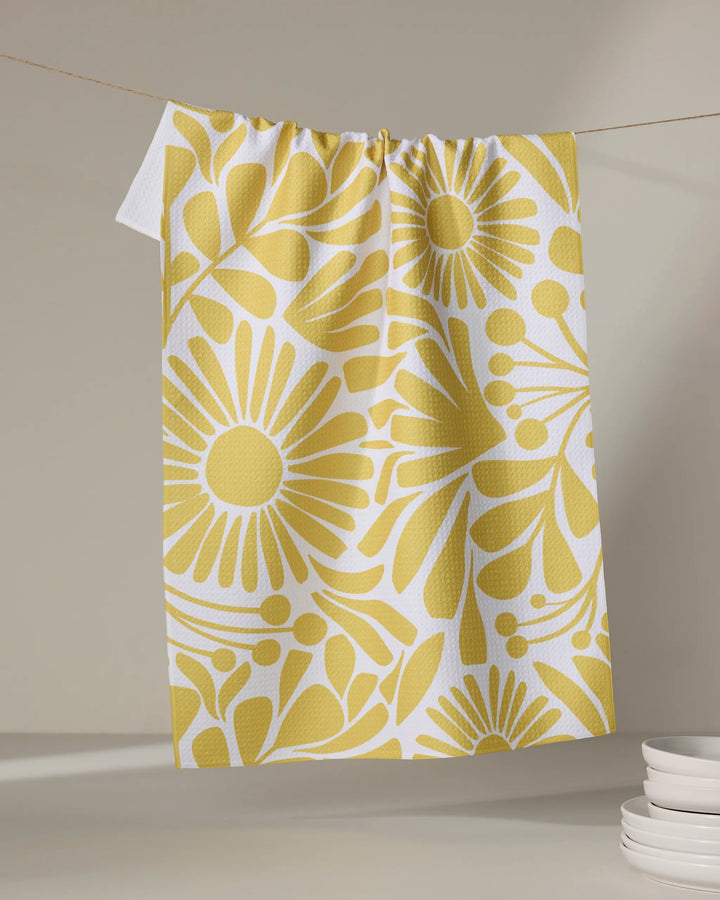 The Sunlit Blooms kitchen towel by Lisa Nelson, made from recycled materials, showcases a bold yellow floral design and hangs on a clothesline. White plates are stacked in the lower right against a neutral backdrop.