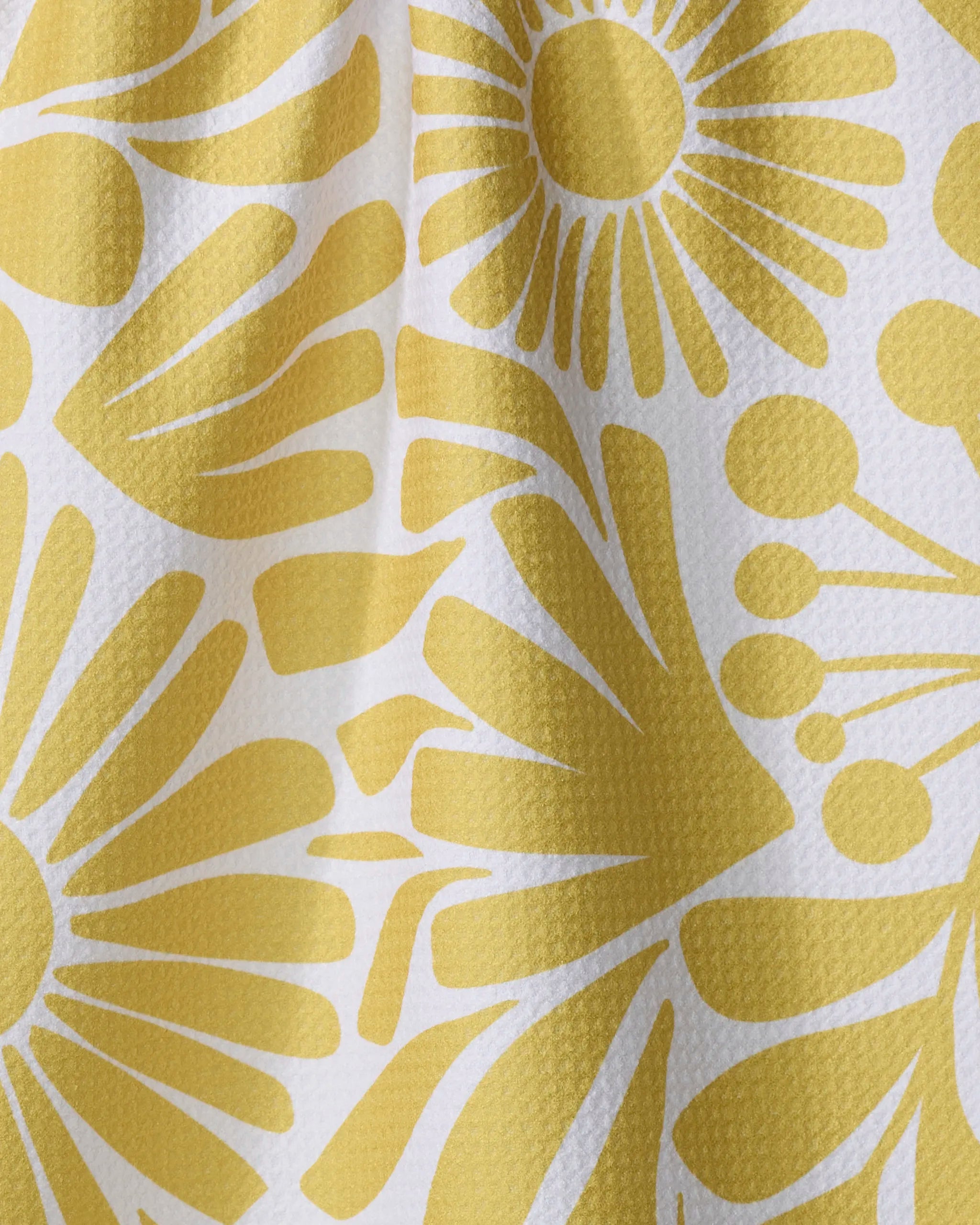 Close-up of Lisa Nelson's Sunlit Blooms kitchen towel, made from textured white recycled material and adorned with a yellow abstract floral pattern featuring stylized flowers and radiating petals, with gentle folds and shadows visible.