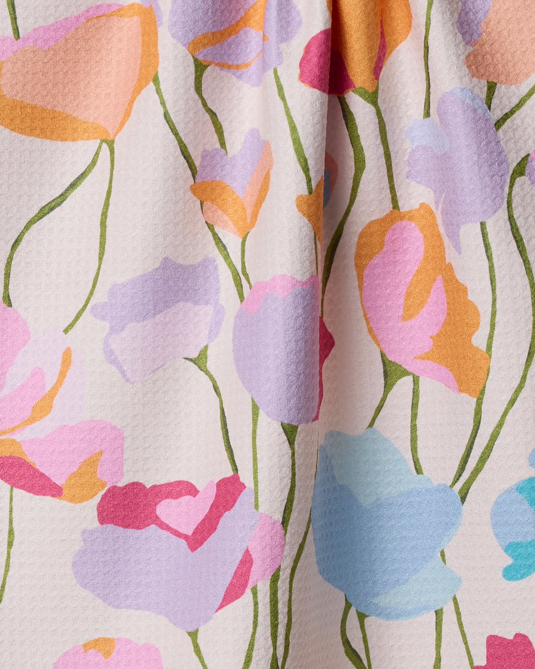 A close-up of the Geometry Sunlit Garden kitchen tea towel, showcasing a pastel floral pattern with abstract pink, orange, purple, and blue flowers on a light background. The slightly textured fabric is softly draped.