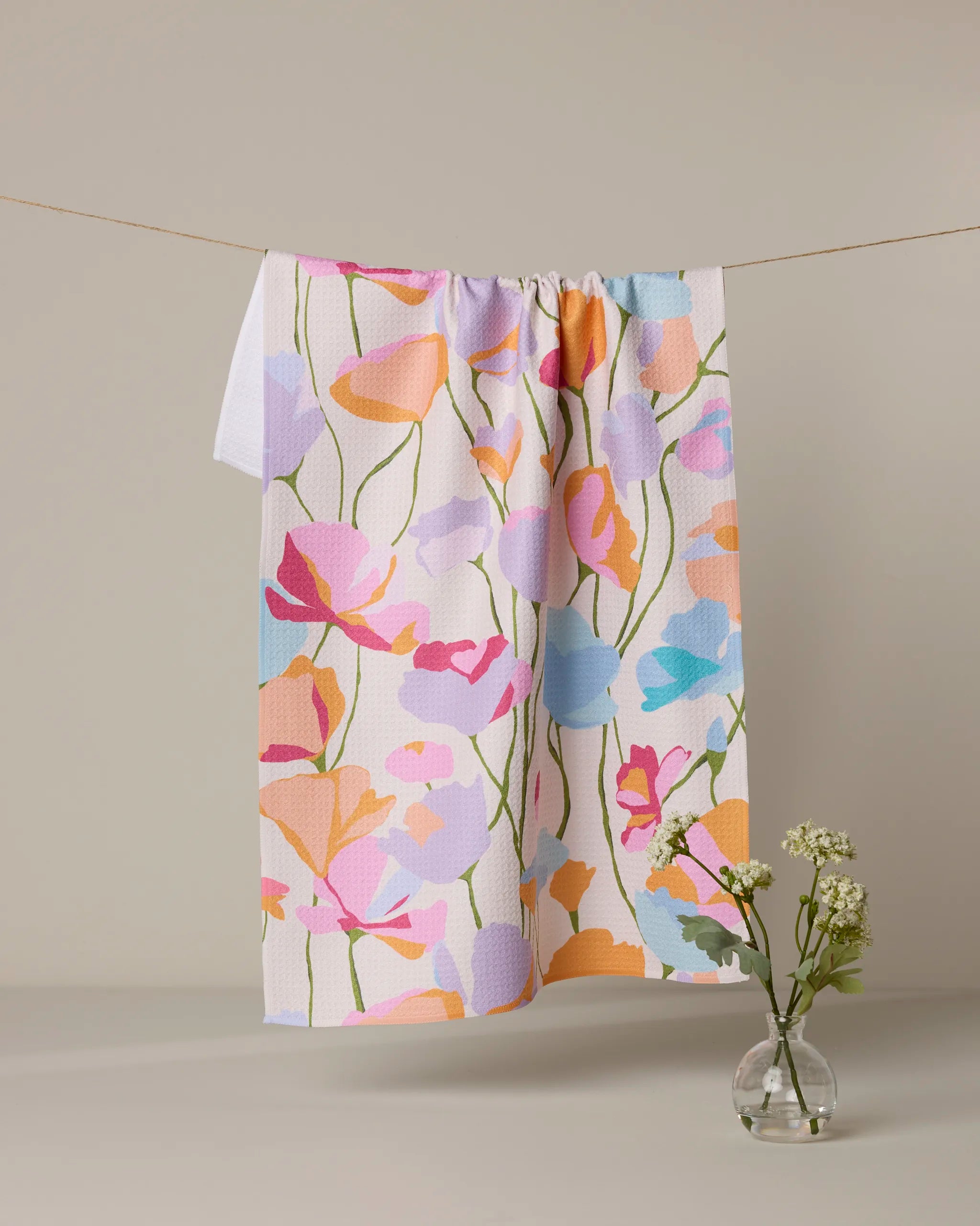 The Geometry Sunlit Garden towel, featuring a vibrant floral pattern and eco-friendly design, hangs on a clothesline with a neutral backdrop. Beneath it sits a small clear vase holding white flowers with green stems.