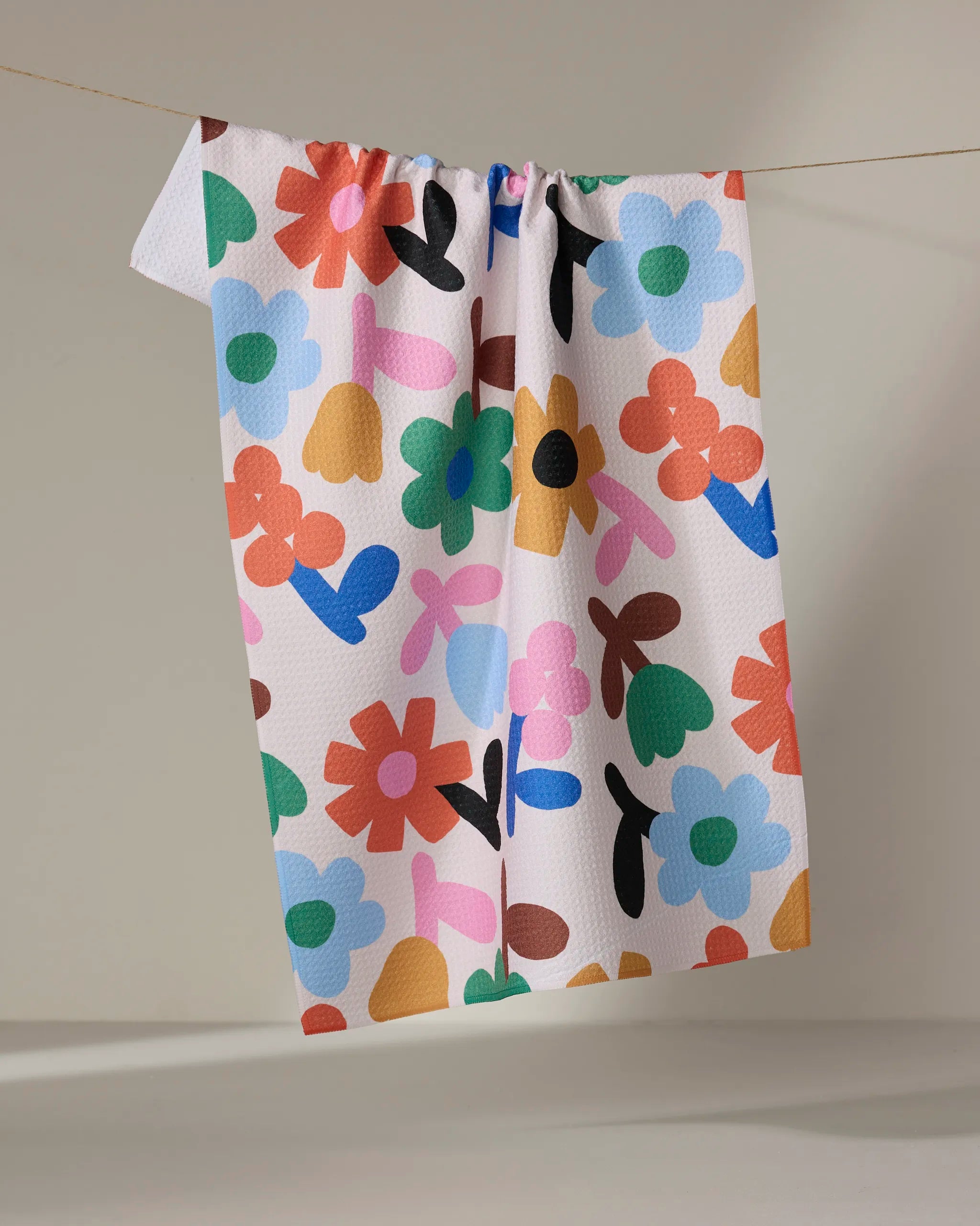 The "Sunny Bloom" kitchen towel by Jennifer Bouron, eco-friendly and white with a vibrant abstract floral pattern in red, blue, green, orange, black, and pink, hangs on a clothesline against a neutral background.