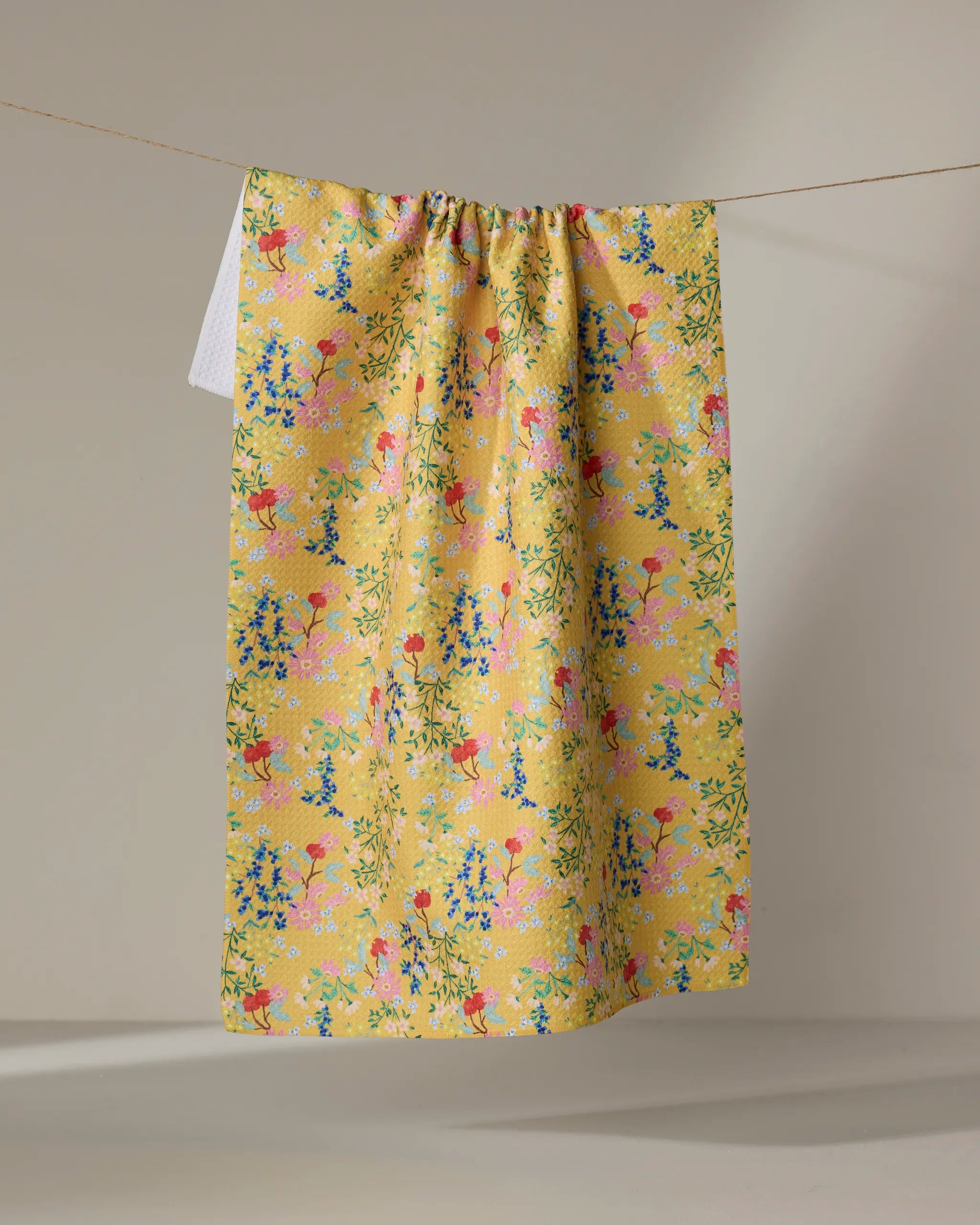 The Brittany Jepsen "Sunny Cherry Blossoms" towel, featuring a colorful floral pattern in yellow, hangs on a string against a plain, light backdrop.