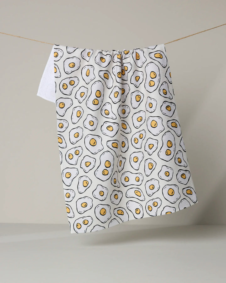 The Rosa Kichline “Sunny Side Eggs” tea towel, made from recycled materials and decorated with a cartoon sunny side up egg pattern, hangs on a clothesline against a neutral background.