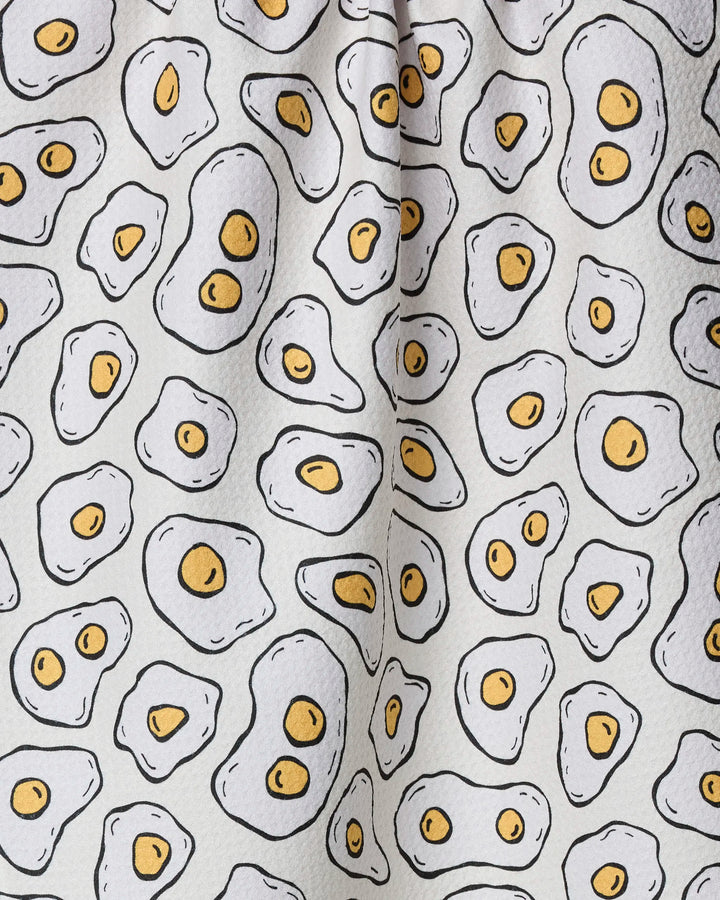 The "Sunny Side Eggs" tea towel by Rosa Kichline is made from recycled materials and showcases a repeating cartoon fried egg pattern—yellow yolks with irregular whites on a white background.