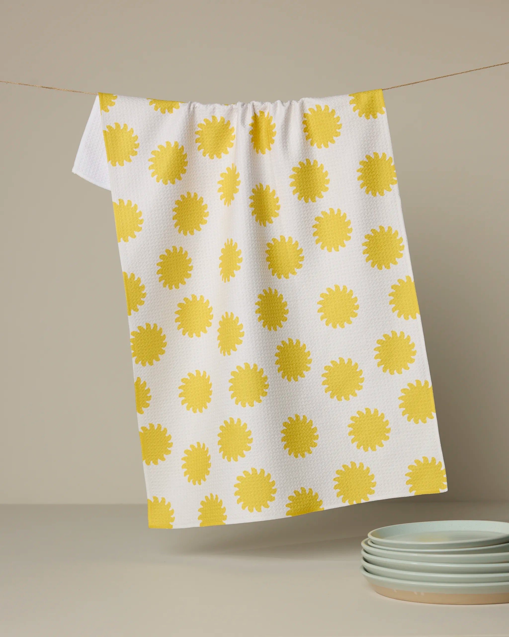 The Sunny Side tea towel by Geometry, featuring bright yellow sunbursts on white and made from recycled materials, hangs on a line with three stacked light blue plates visible in the lower right against a beige background.