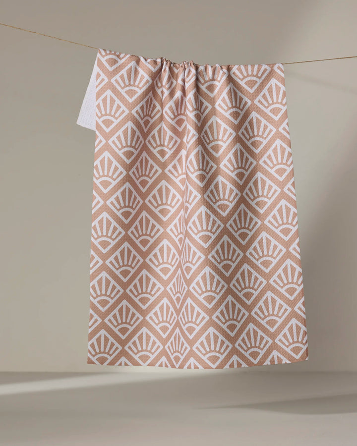 The Rosa Kichline "Sunrise Mosaic" recycled tea towel in blush pink and white features a geometric fan pattern and hangs from a string against a neutral beige background.
