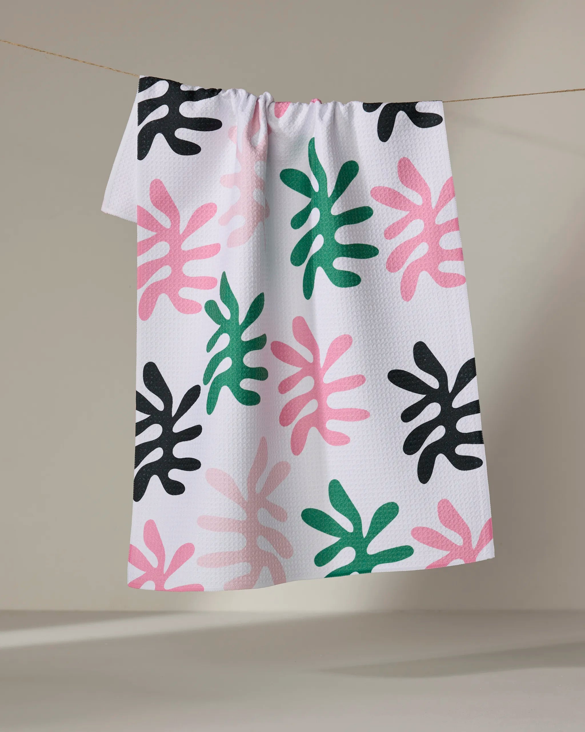 The Marina Solodka "Sunrise" tea towel, featuring abstract leaf-like shapes in black, pink, and green, hangs against a plain light background.
