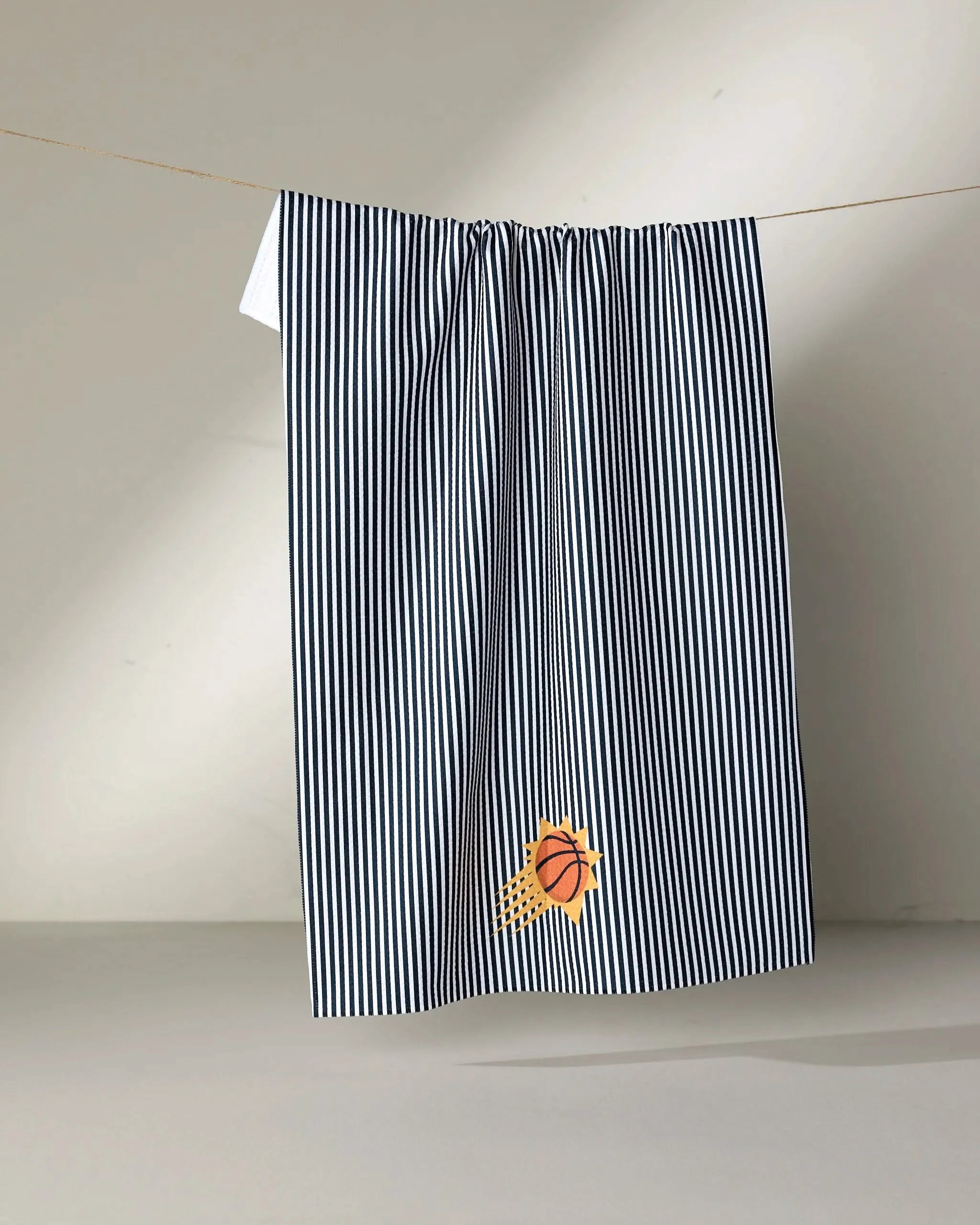 A black-and-white striped NBALAB x Geometry "Suns Classic" NBA towel hangs on a line. A small embroidered basketball with a yellow splash accents the bottom corner. The background is plain and light-colored.