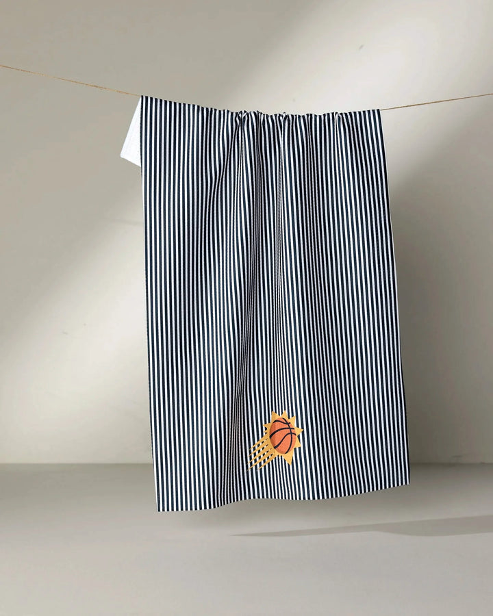 A black-and-white striped NBALAB x Geometry "Suns Classic" NBA towel hangs on a line. A small embroidered basketball with a yellow splash accents the bottom corner. The background is plain and light-colored.