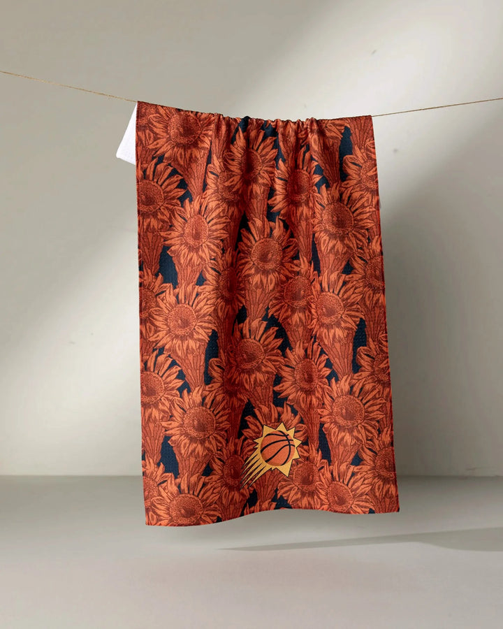 The Suns Floral tea towel by NBALAB x Geometry features a reddish-orange sunflower pattern with a small Phoenix Suns basketball logo near the bottom, hanging on a string against a neutral background. Part of the NBA tea towel collection.